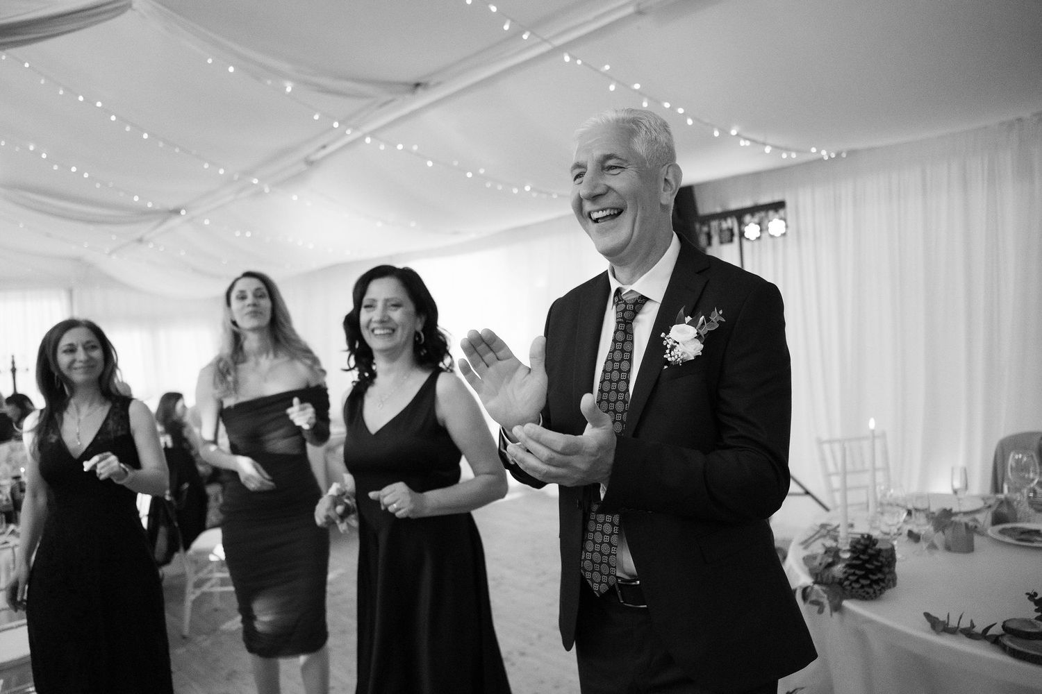 Black and white photo of wedding guests clapping and laughing joyfully inside an elegant tent reception venue.