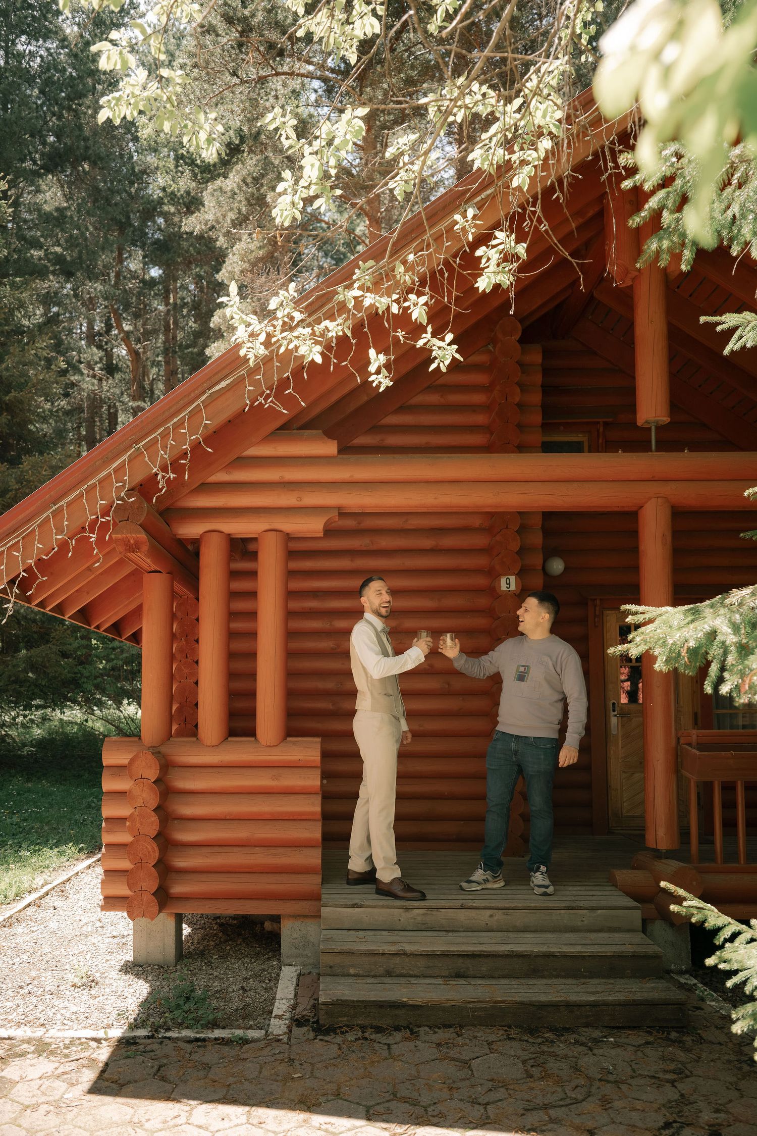 Two men shaking hands on the porch of a rustic log cabin surrounded by tall trees in a forest setting.