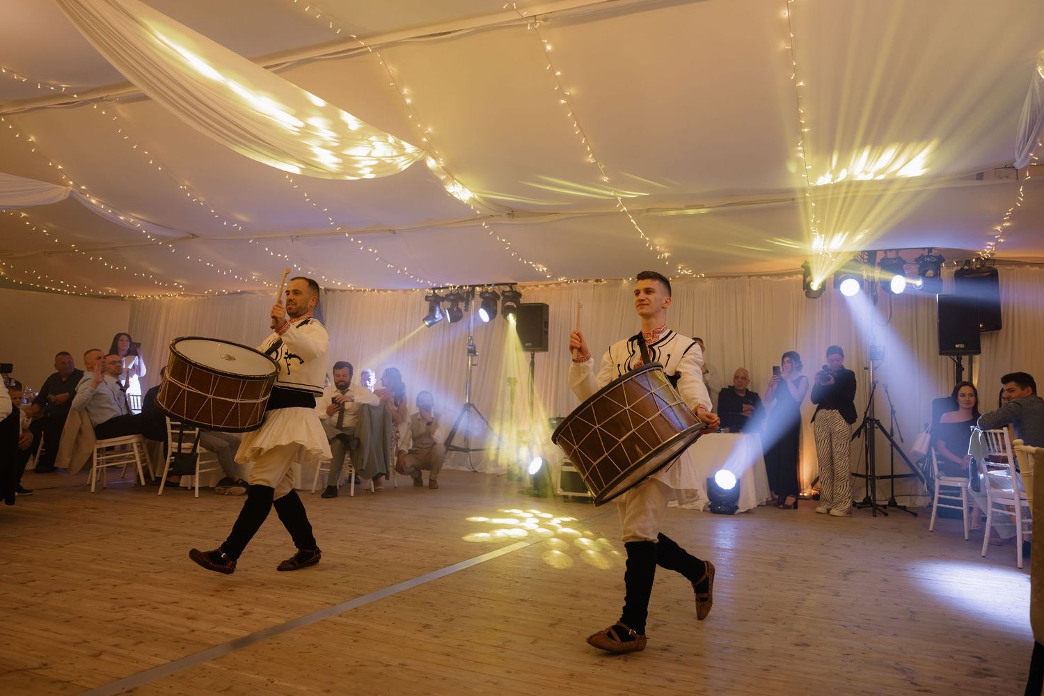 Two drummers in traditional costumes perform at an elegant wedding reception with fairy lights and stage lighting.
