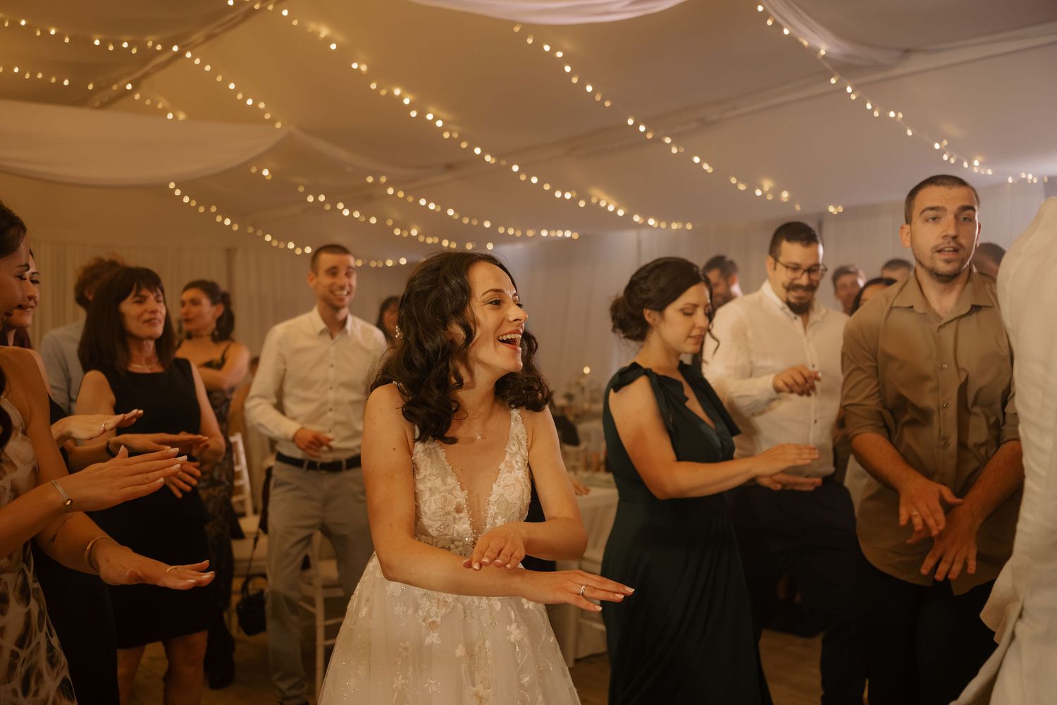 Bride in white gown dancing joyfully with guests at elegant wedding reception with fairy lights.