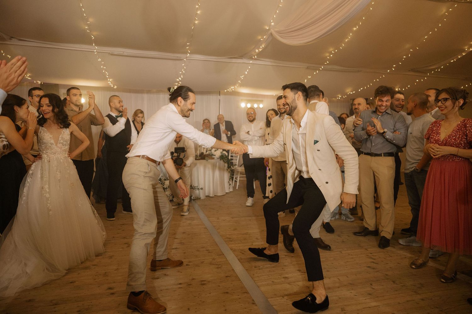 Two grooms dancing joyfully at their wedding reception surrounded by cheering guests in a decorated tent venue.