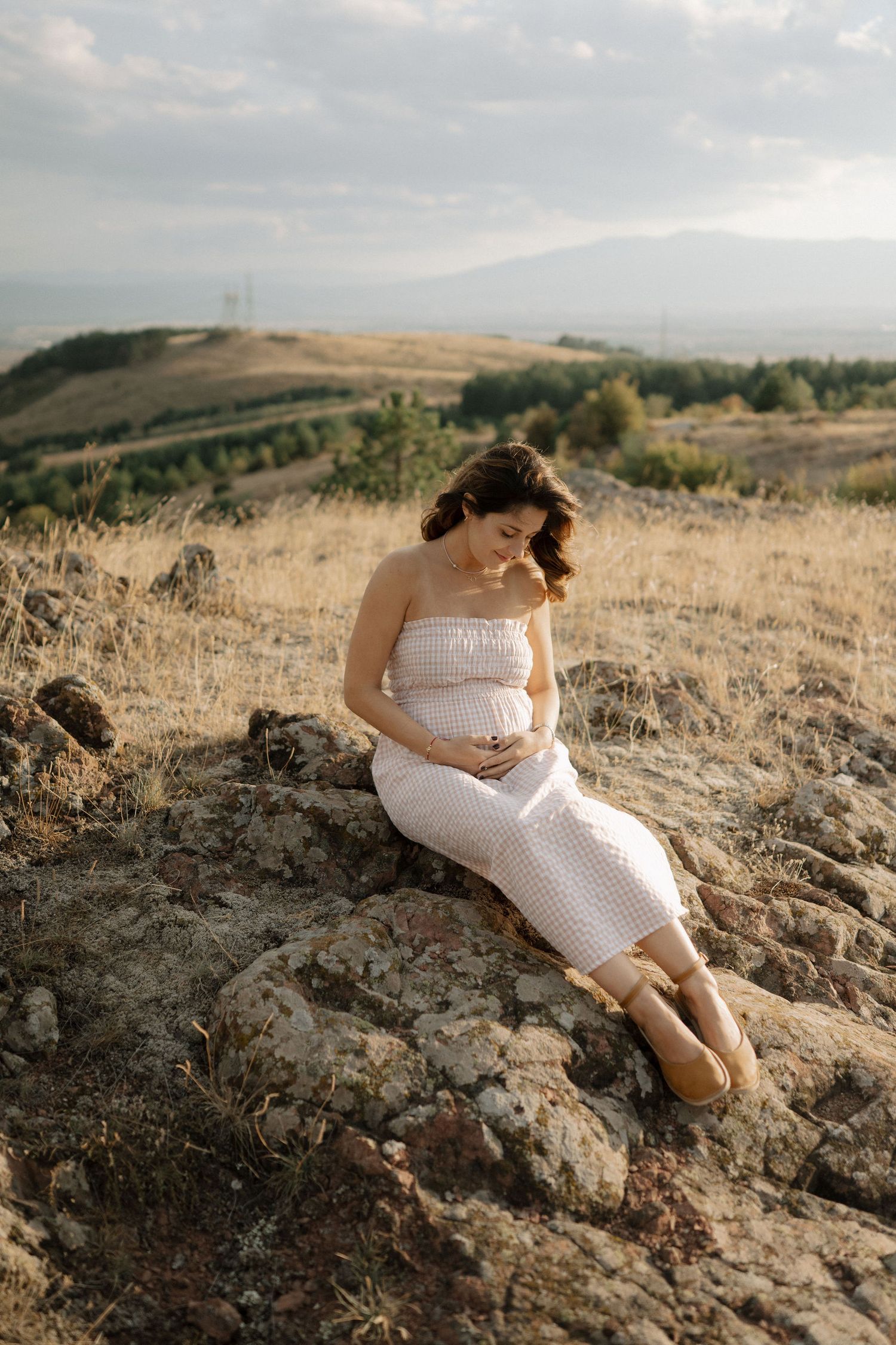A figure in a flowing white dress reclines on rocky terrain against a scenic mountain landscape at golden hour.
