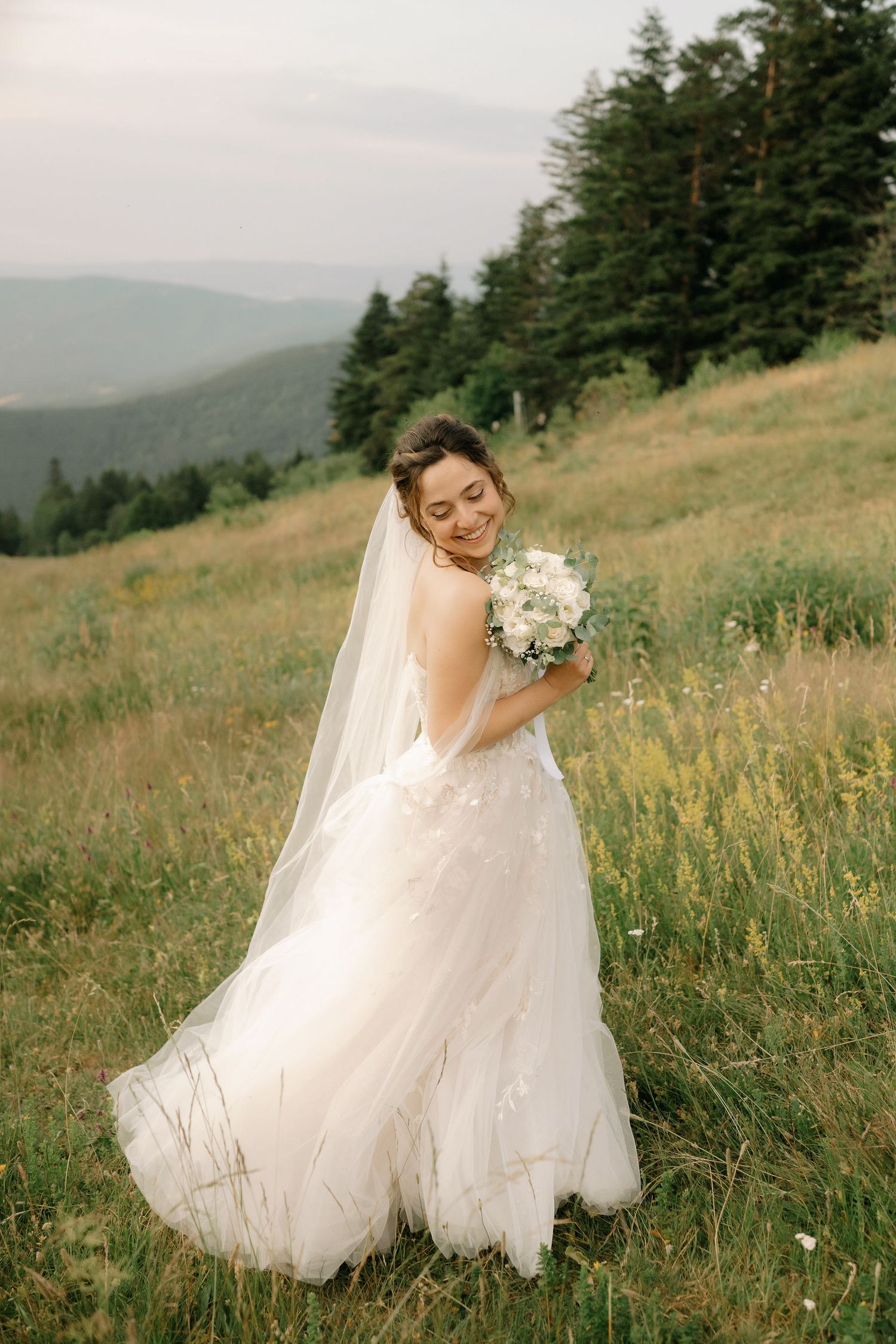 A bride in a flowing white wedding dress twirls gracefully in a mountain meadow at sunset with evergreen trees in background.