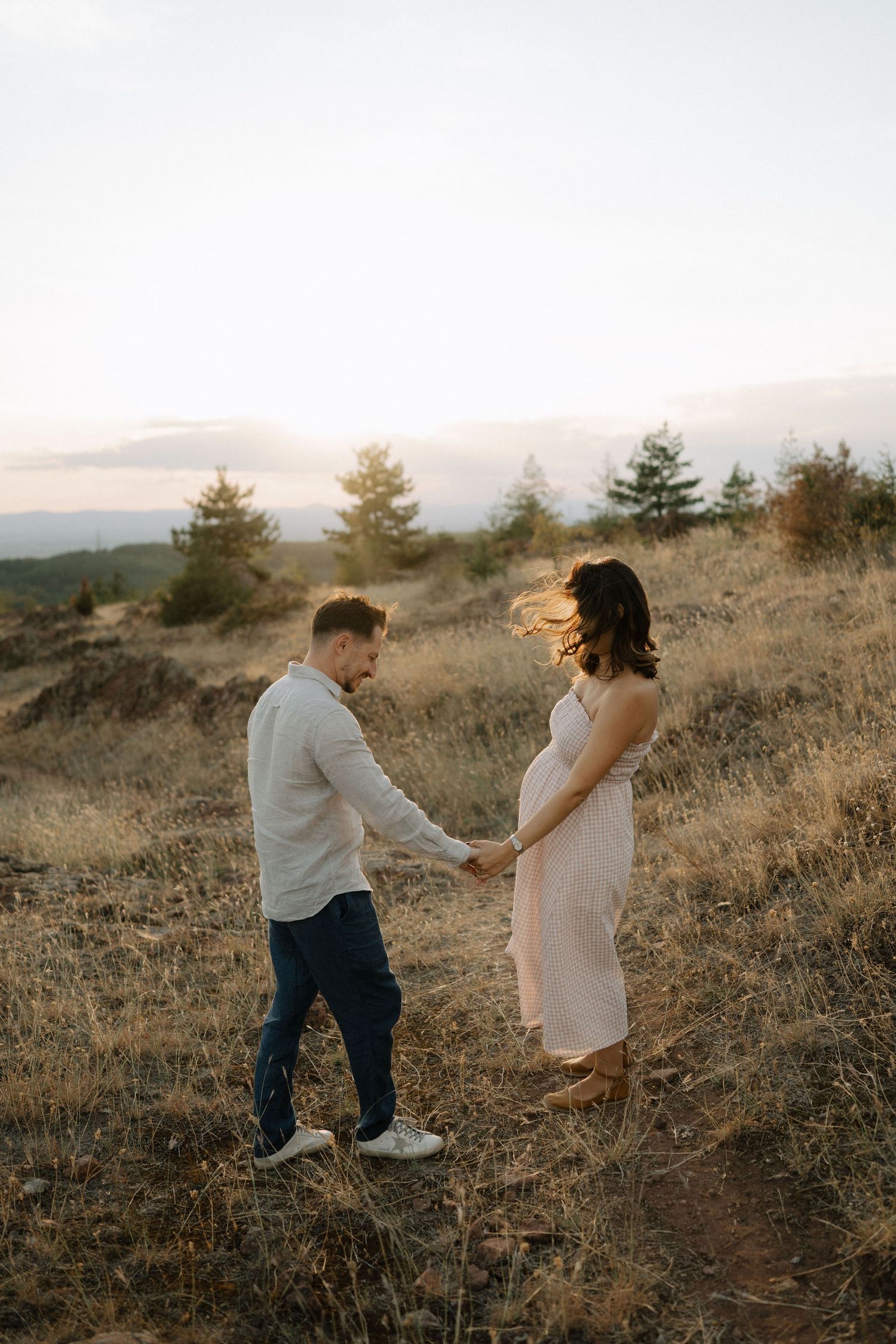 A couple holds hands on a hillside at sunset with evergreen trees and mountains in the background.