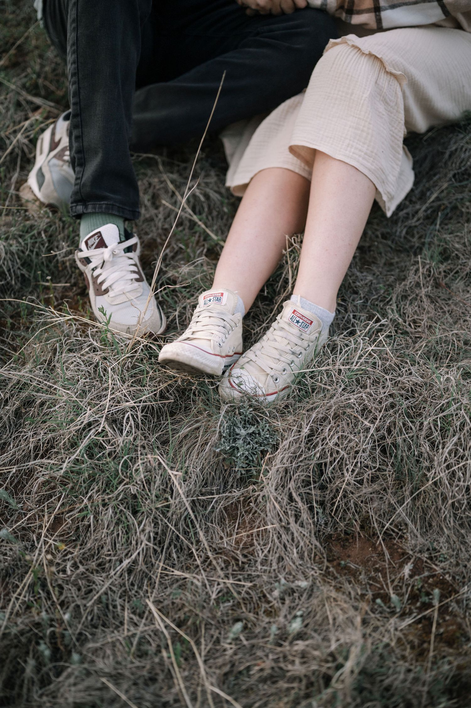 Two pairs of worn white sneakers resting on dry grass with one person wearing khaki pants and another in dark pants.