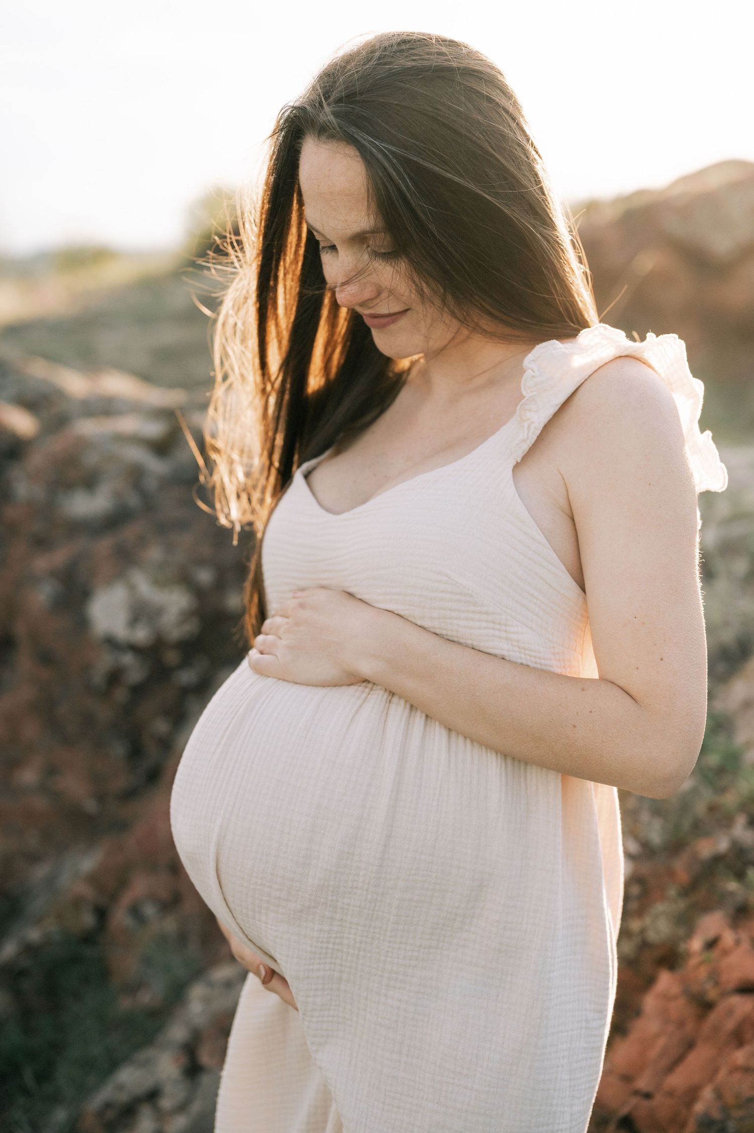 A pregnant woman in a flowing white dress stands against a rocky backdrop during golden hour lighting.