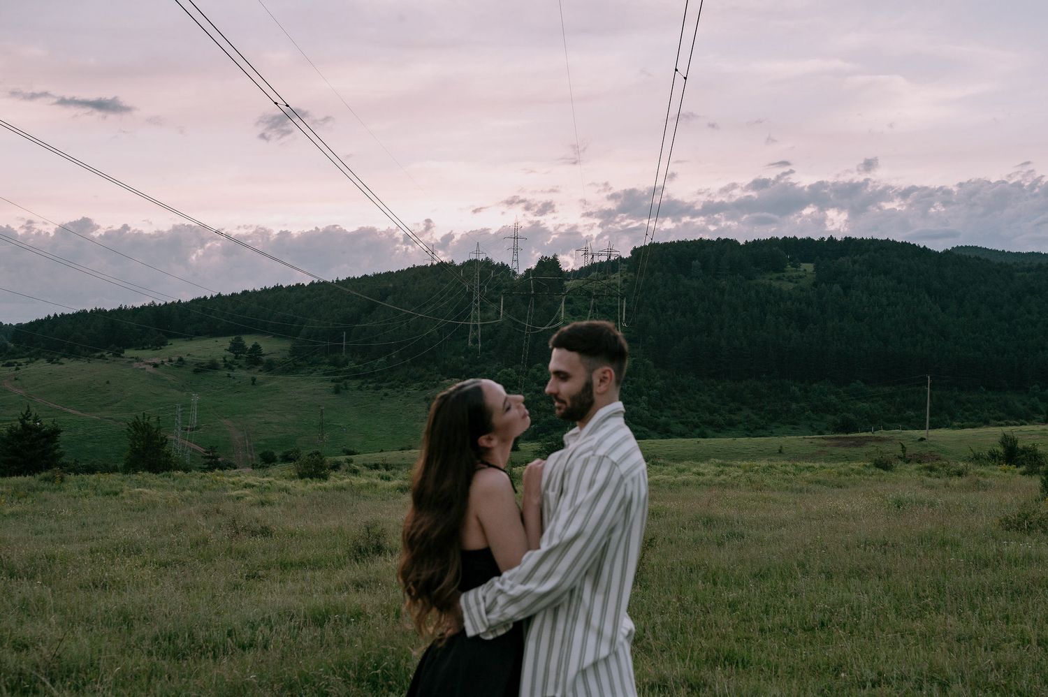 A couple embraces in a romantic moment against a backdrop of rolling green hills and mountains at sunset.