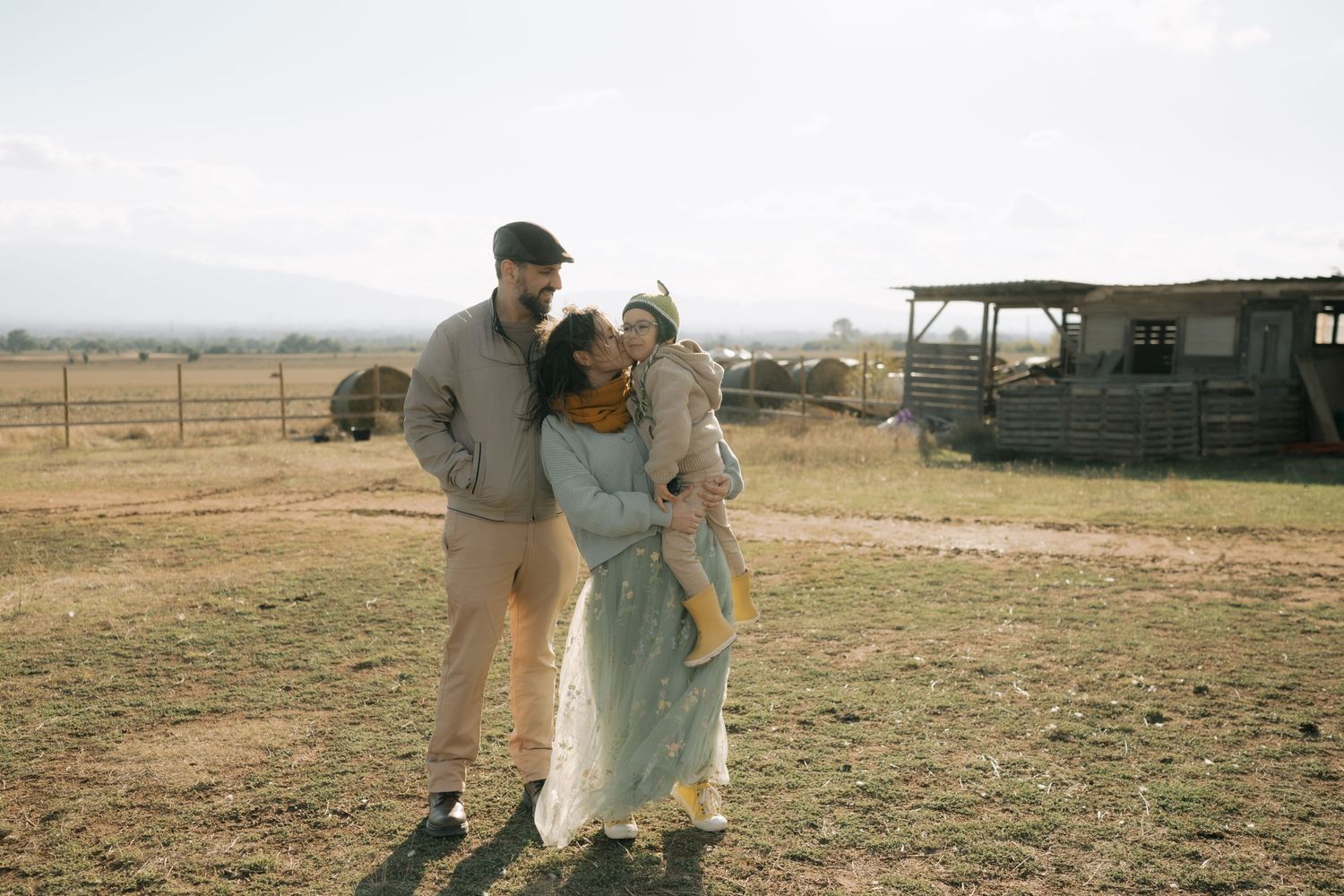 A series of people walking together through a rural farm landscape with barren fields and buildings in the distance.
