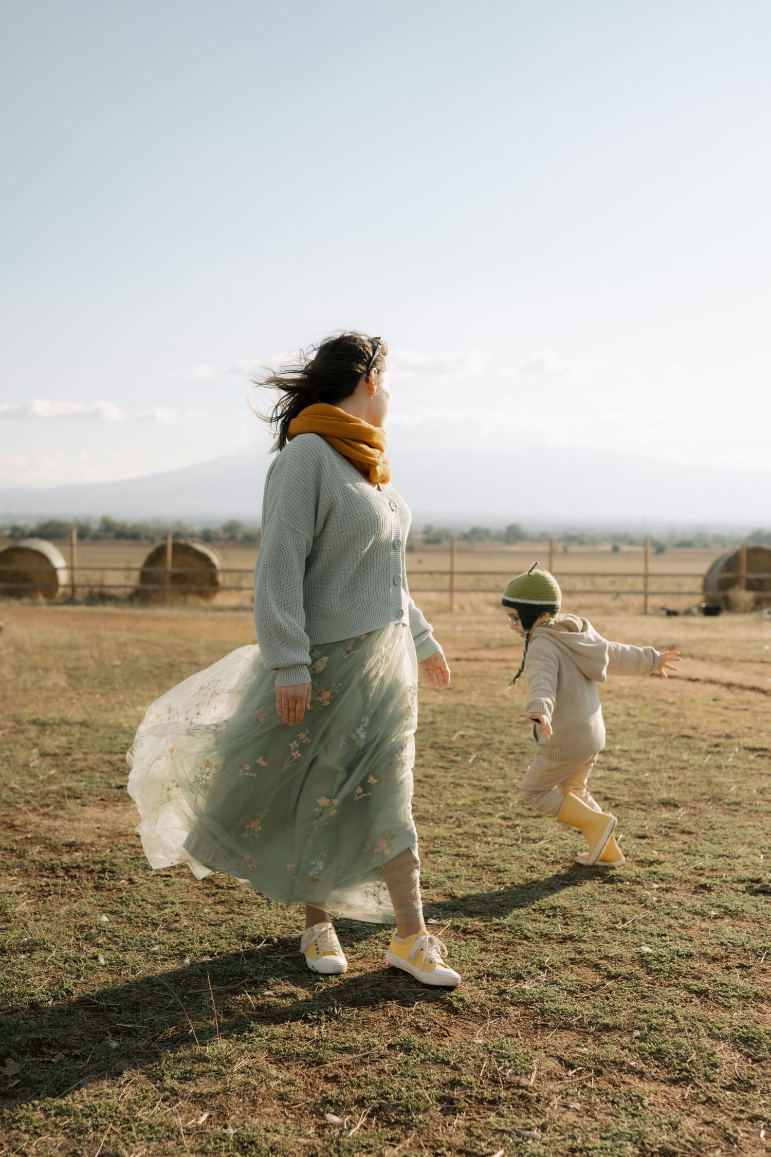 A figure in a flowy dress runs through a sunlit field with a white dog companion on a golden afternoon.