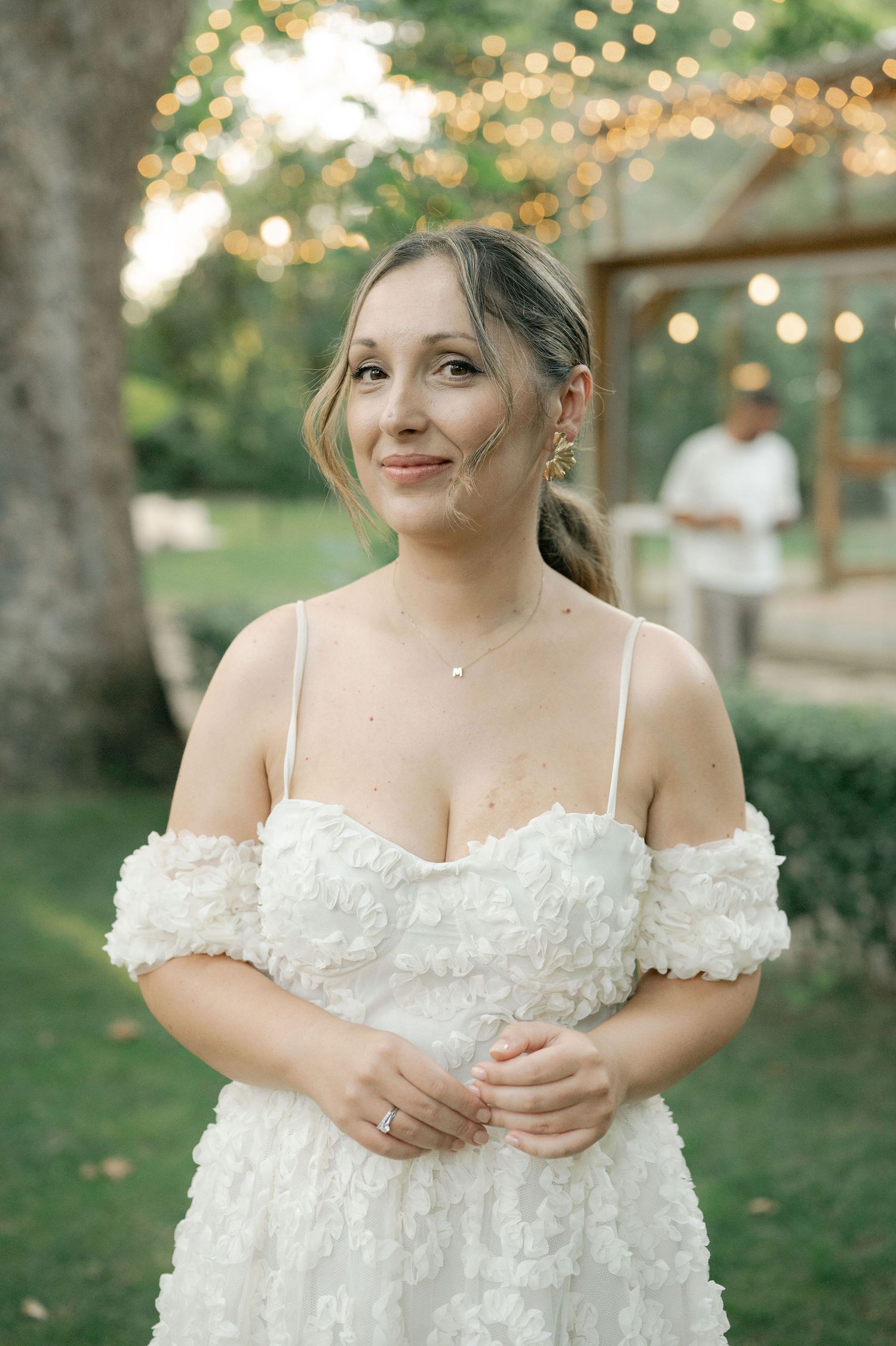 Bride in off-shoulder white wedding dress with delicate floral details stands in outdoor garden setting with bokeh lights.