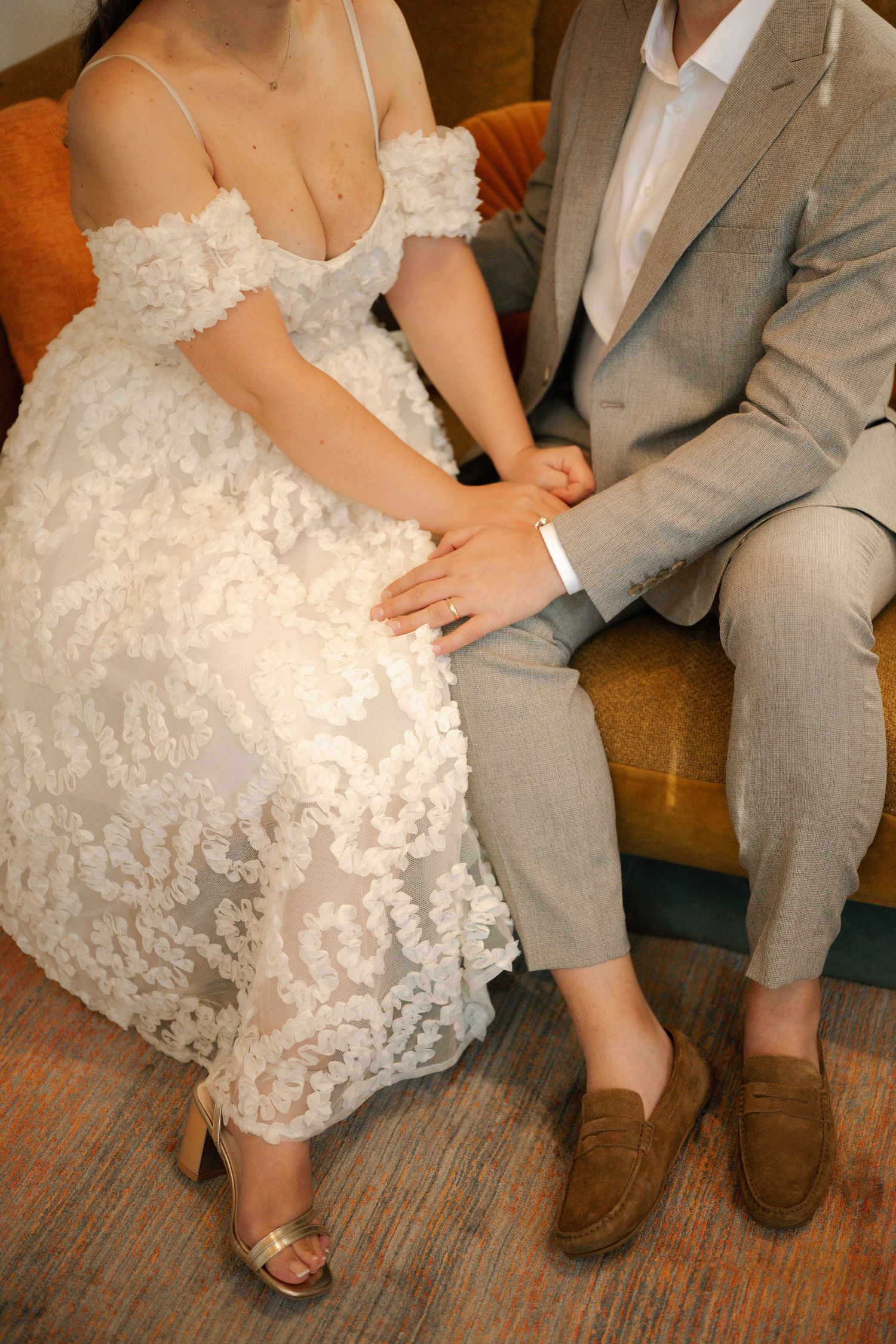 Close-up details of a bride in white lace dress and groom in gray suit sitting together showing their shoes.