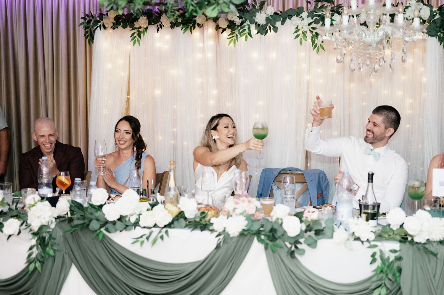 Wedding reception table decorated with elegant white linens, floral arrangements and crystal chandeliers in a romantic setting.