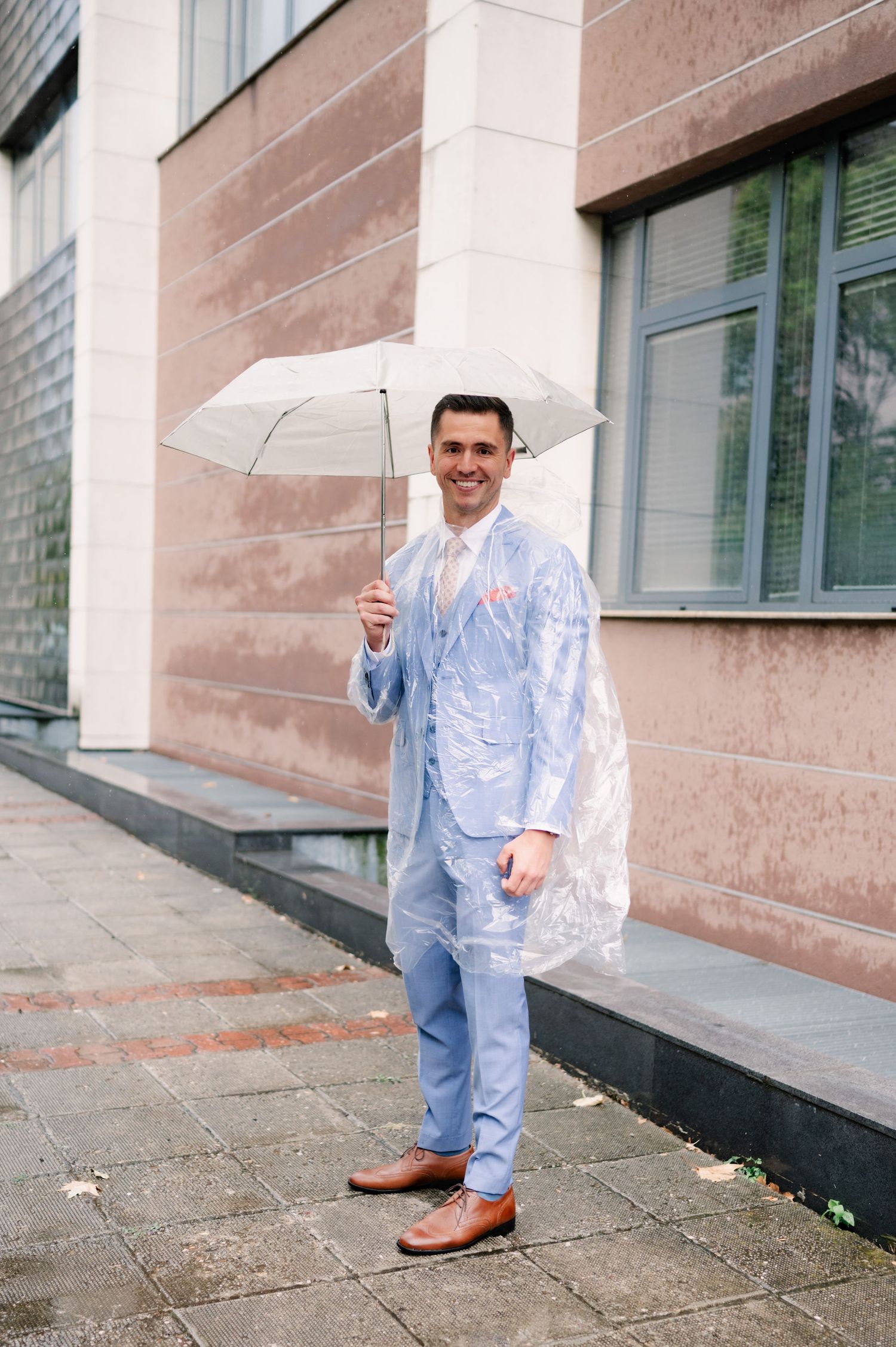 A person in a light blue outfit holds a clear umbrella while standing outside a building with pink stone walls.