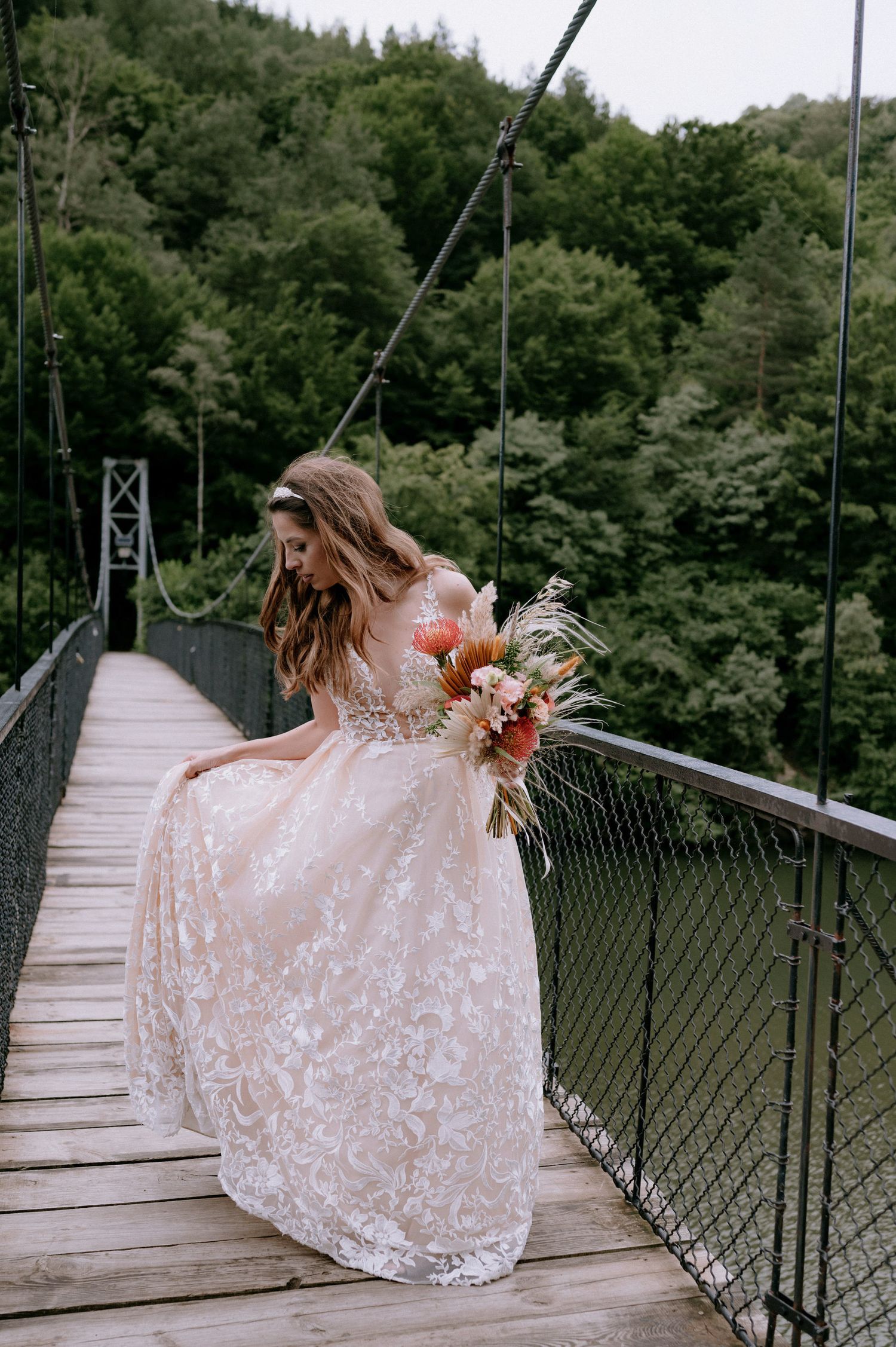 A person in a flowing white lace dress poses gracefully on a scenic suspension bridge surrounded by lush forest.
