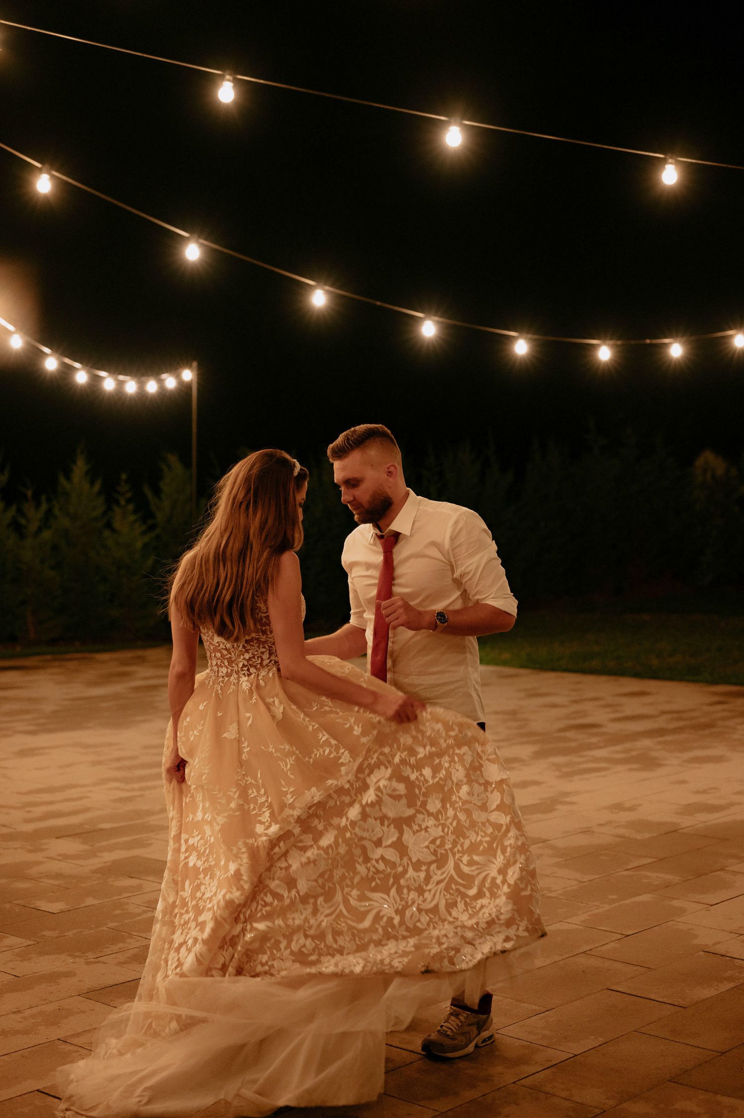A couple dances together under warm string lights on an outdoor patio during an evening celebration.