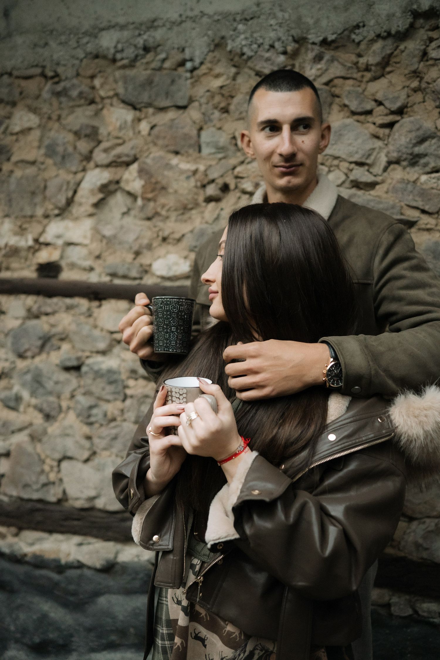 Couple enjoying hot beverages together while leaning against a rustic stone wall.