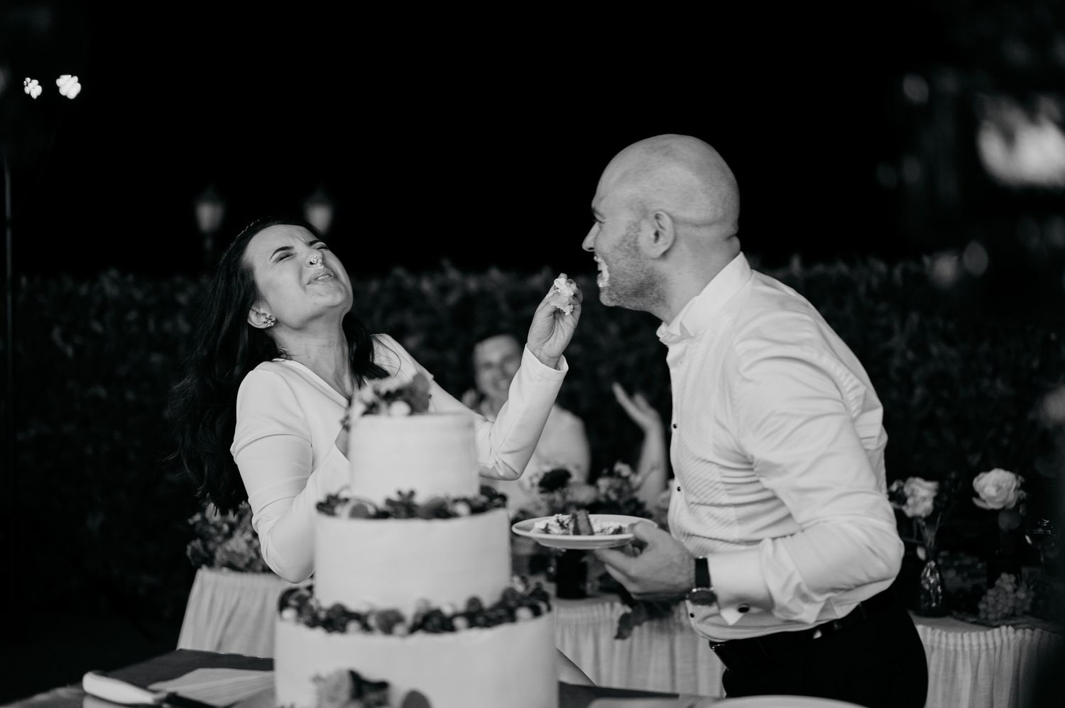A couple shares a playful moment while feeding each other wedding cake at their reception in black and white.