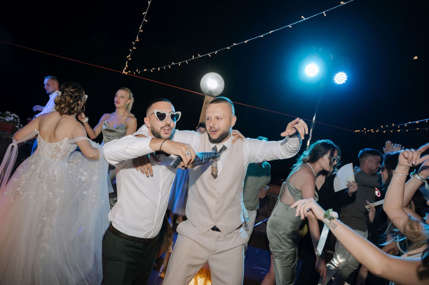 Wedding guests dancing together under blue lighting and string lights at an outdoor nighttime reception celebration.