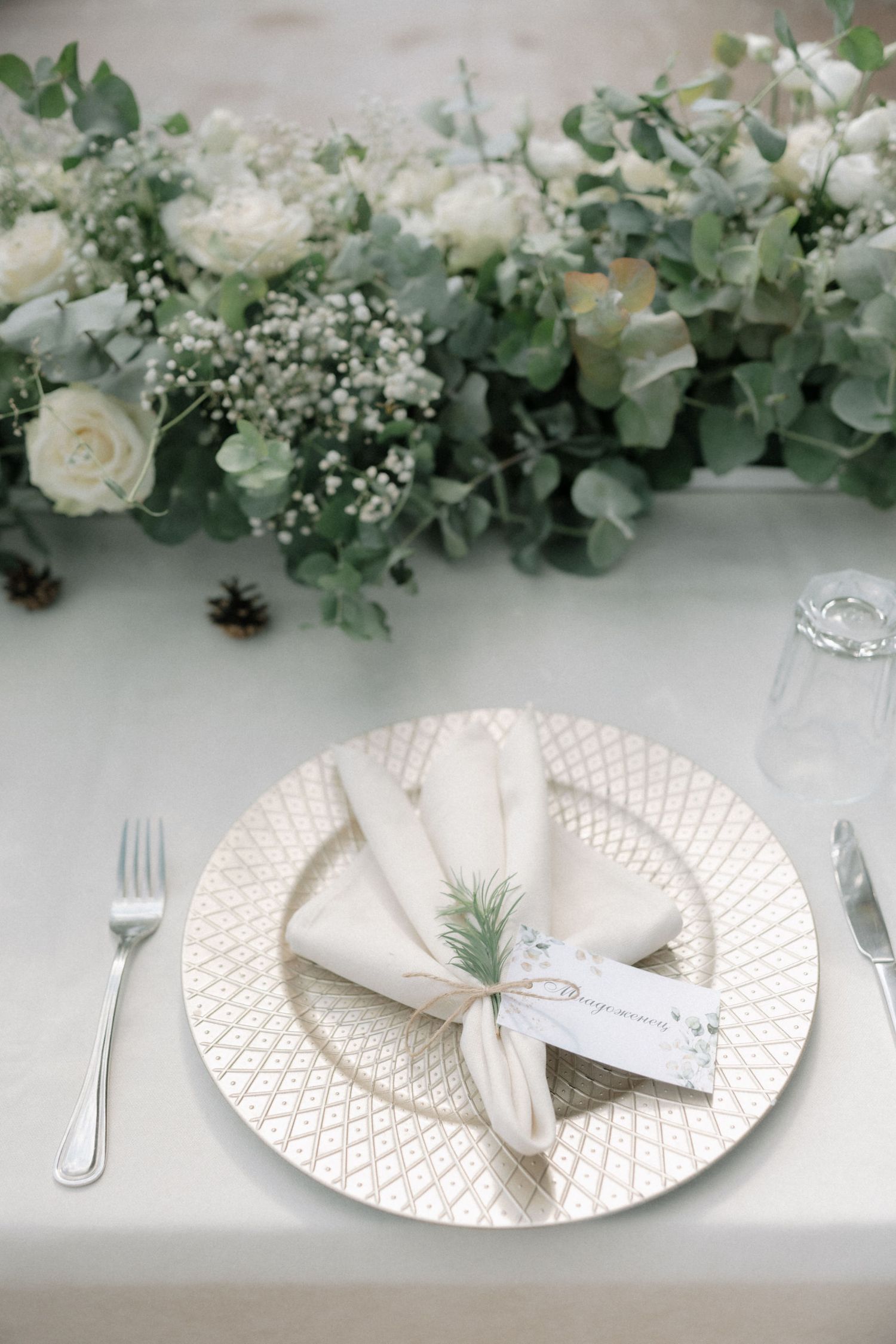 Elegant white and gold place settings with eucalyptus garland and baby's breath floral arrangements on a light gray table.