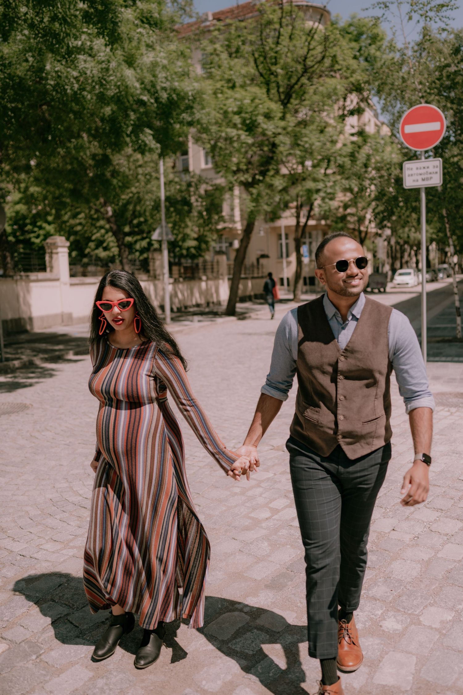 Stylish couple holding hands walking on a sunny cobblestone street, woman in striped maxi dress, man in vest and sunglasses.