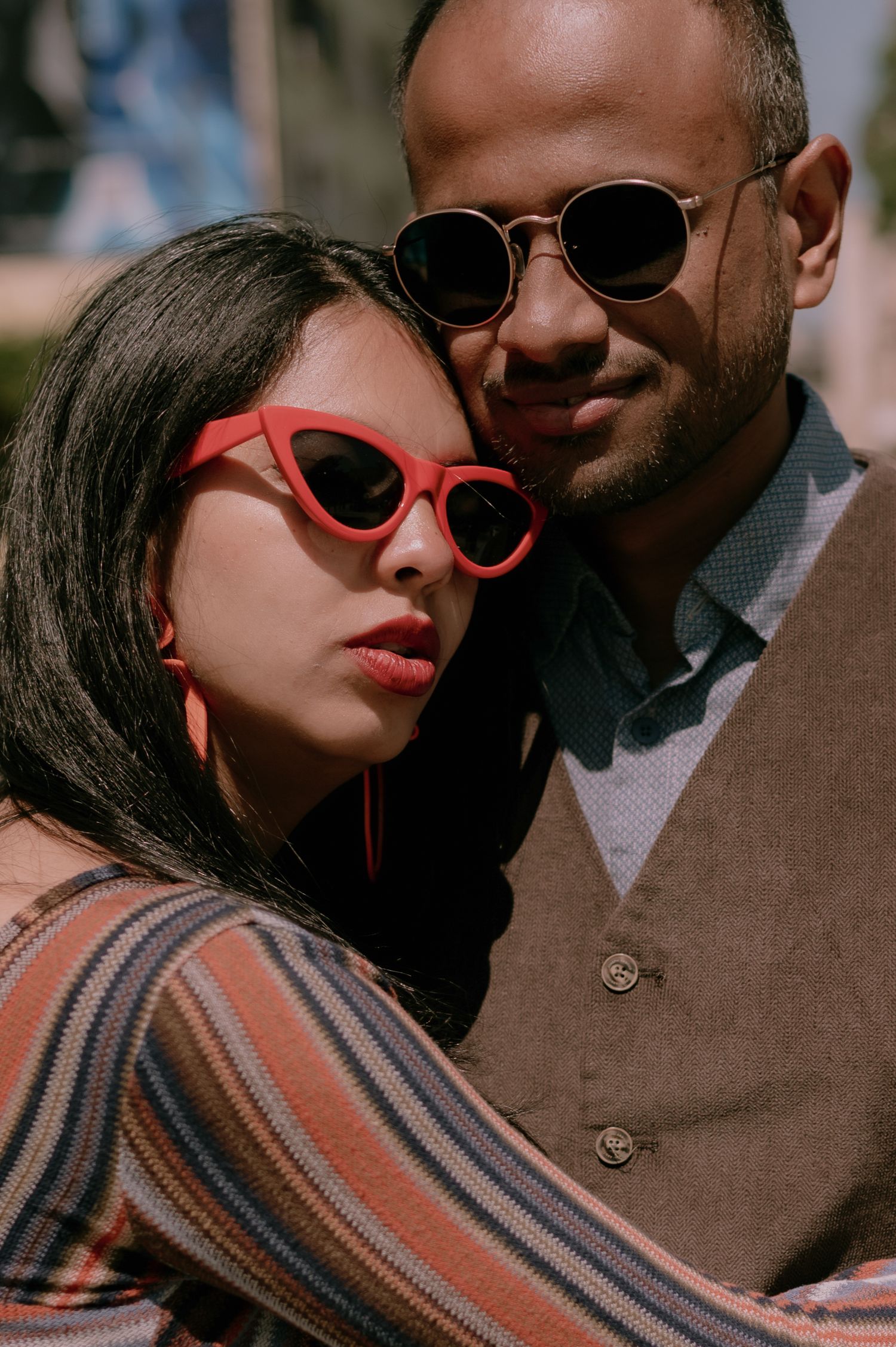 A stylish couple wearing sunglasses pose closely together, woman in red cat-eye frames and striped top, man in gold aviators.