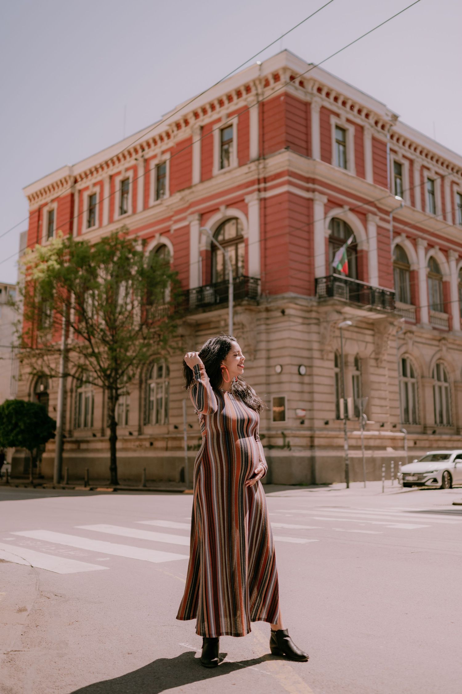 Pregnant woman in striped maxi dress poses on sunny street corner near red historic building.