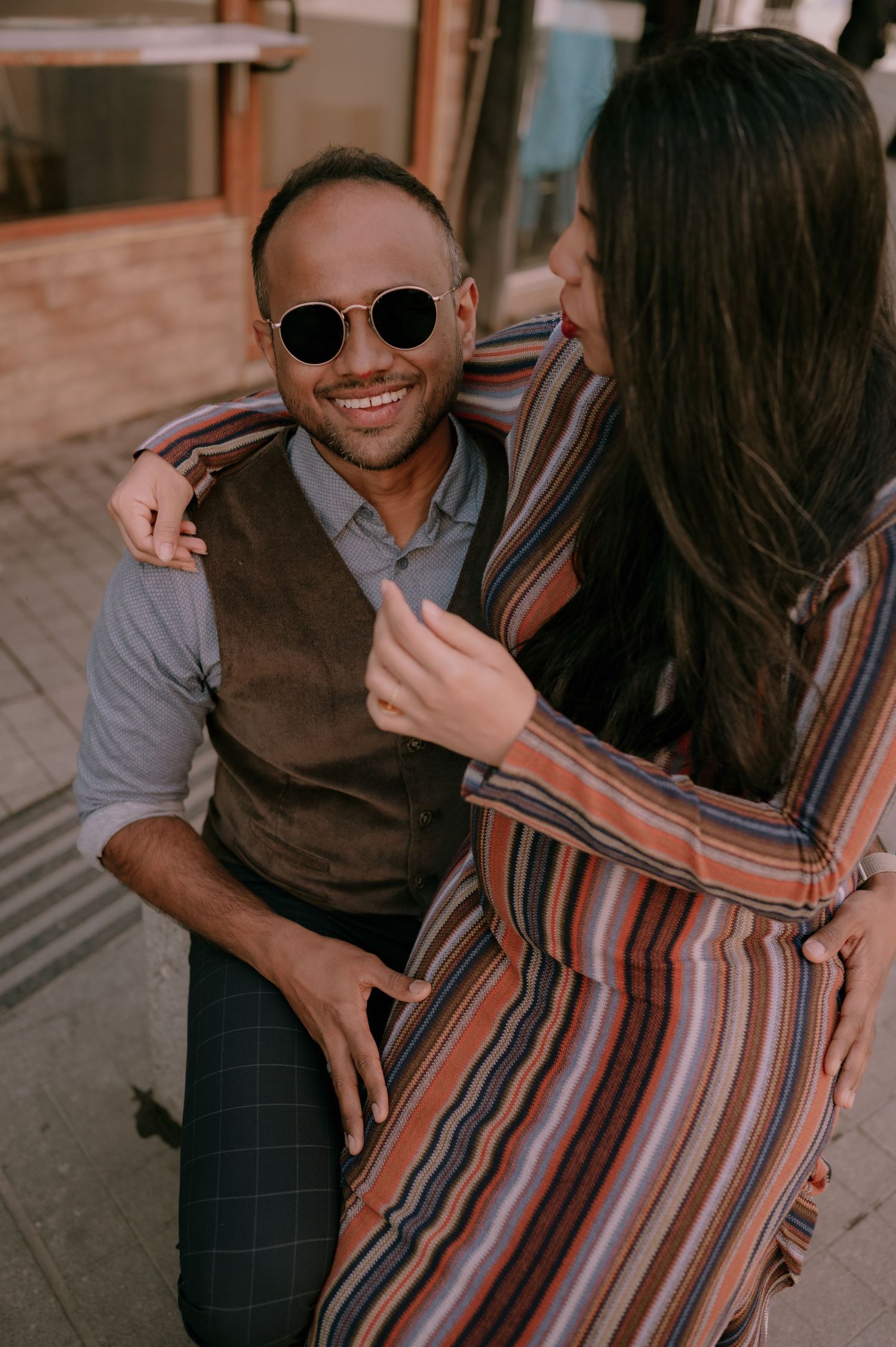A smiling man in sunglasses and vest sits outdoors as a woman in a striped dress affectionately embraces him.