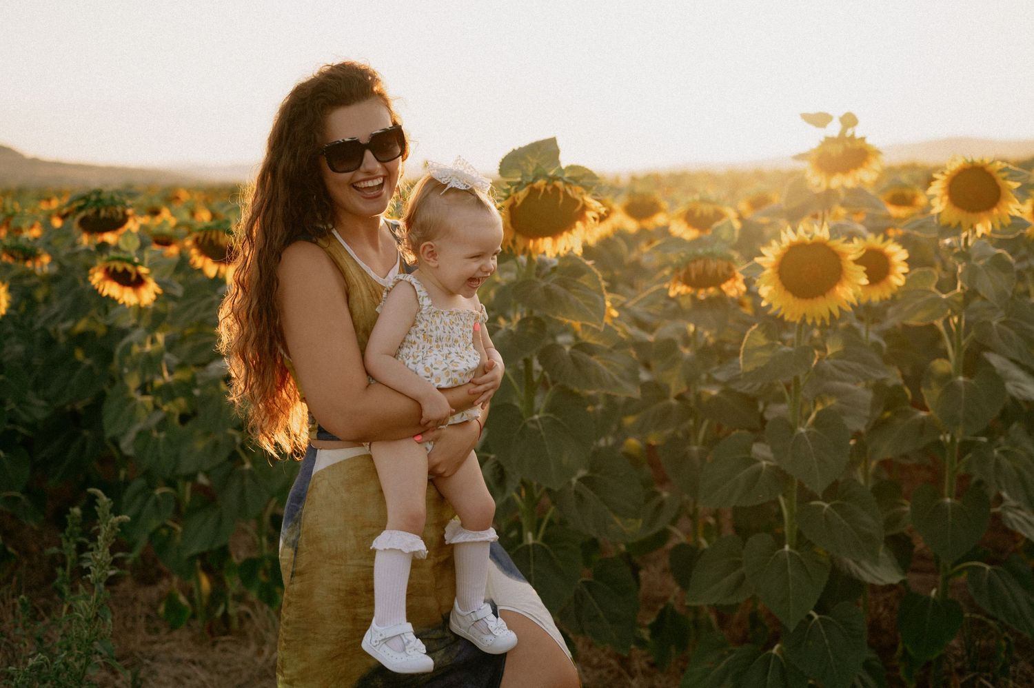 A mother holds her baby girl in a sunflower field at golden hour sunset, both smiling joyfully.