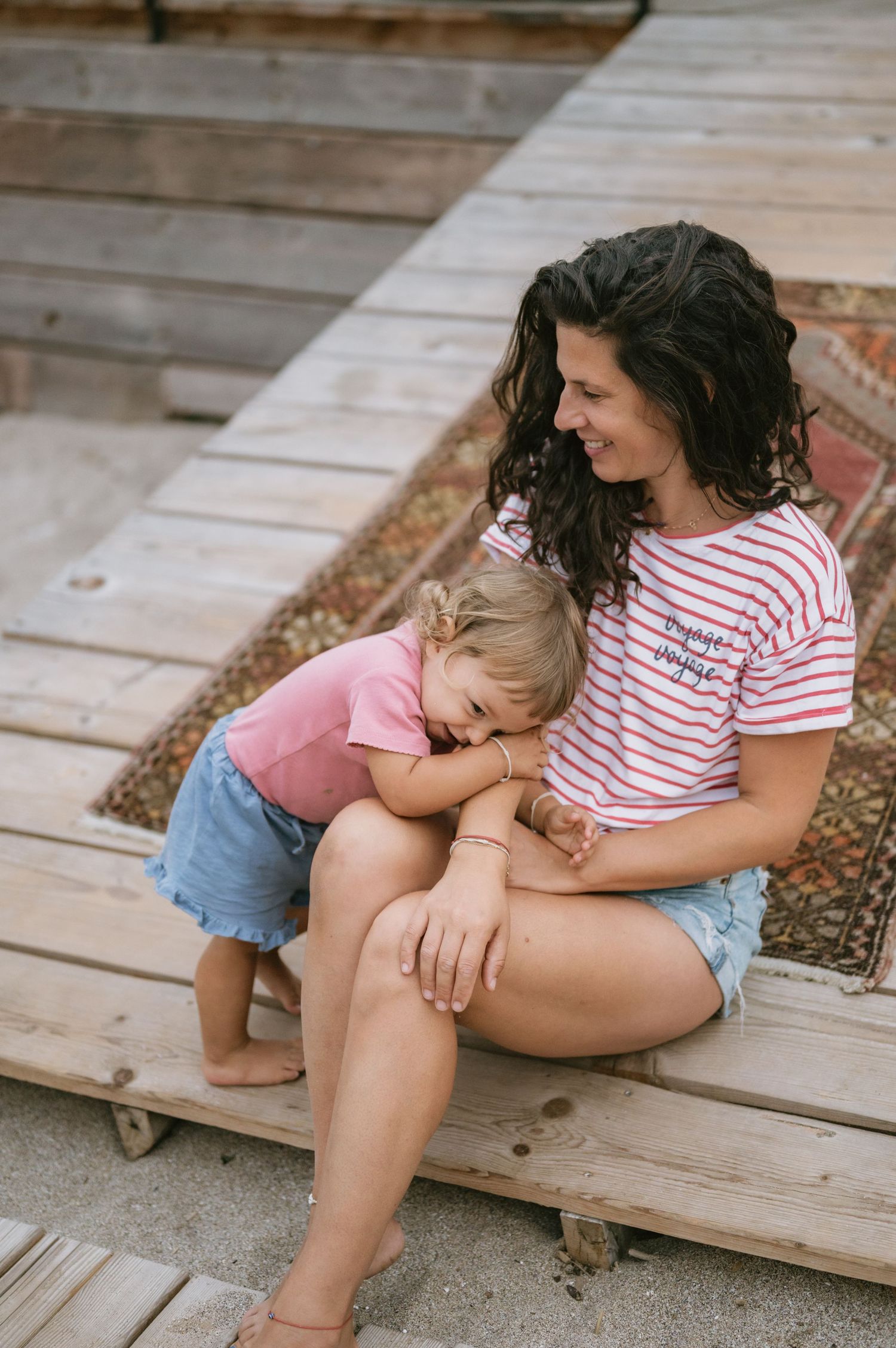 A smiling mother in a striped shirt sits on wooden steps with her toddler daughter leaning against her affectionately.