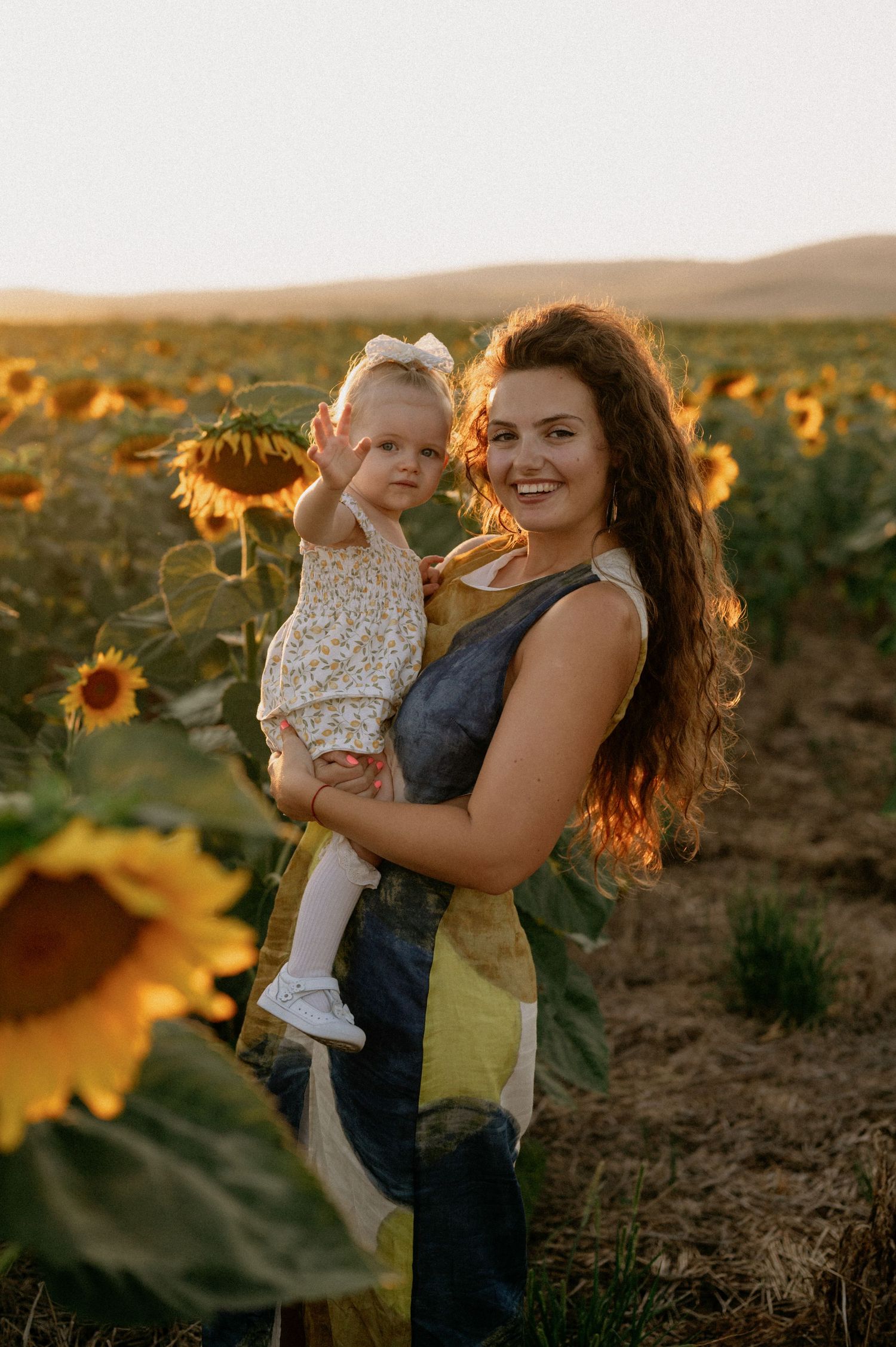 A smiling woman holds a baby girl in a sunflower field during golden hour sunset.