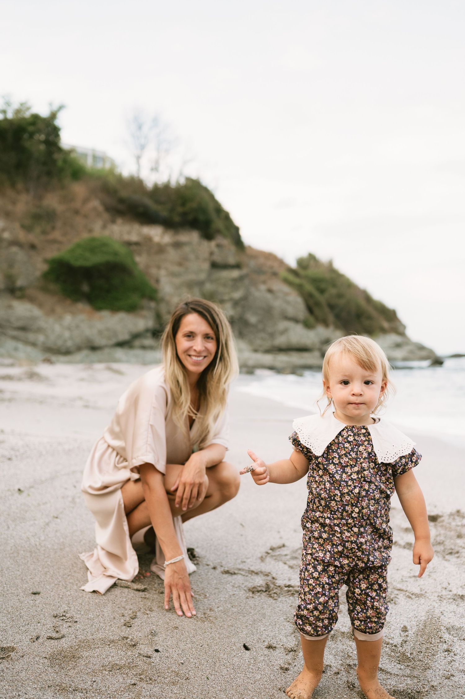 A smiling woman crouches on a sandy beach beside a toddler in a floral romper, with rocky cliffs in the background.