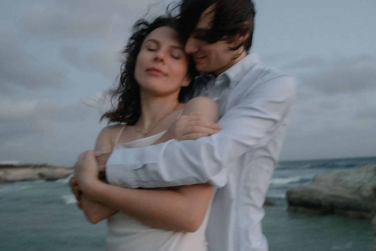 A couple embraces tenderly on a rocky ocean shore, the woman in white with eyes closed, man in blue shirt behind her.