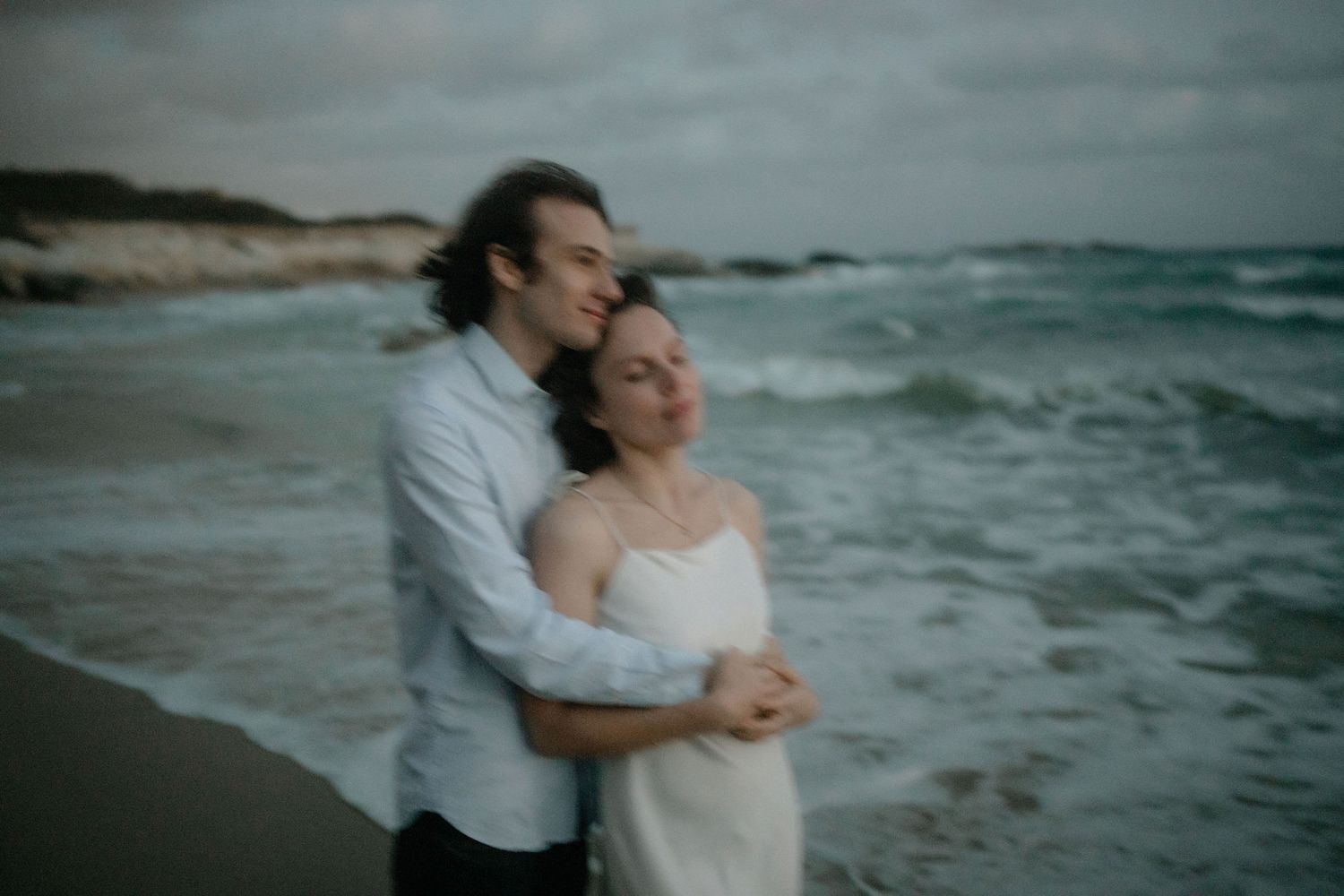 Couple embracing romantically on a beach with ocean waves, man in blue shirt hugging woman in white dress.