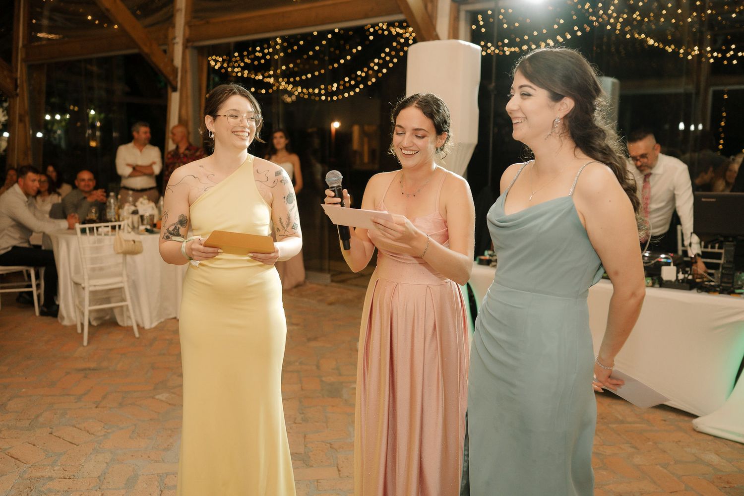 Three bridesmaids in pastel evening gowns chat during a reception under string lights at an indoor venue.