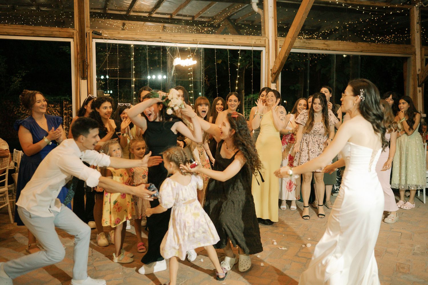A lively wedding reception dance party with guests dancing in a covered outdoor venue under warm lighting.