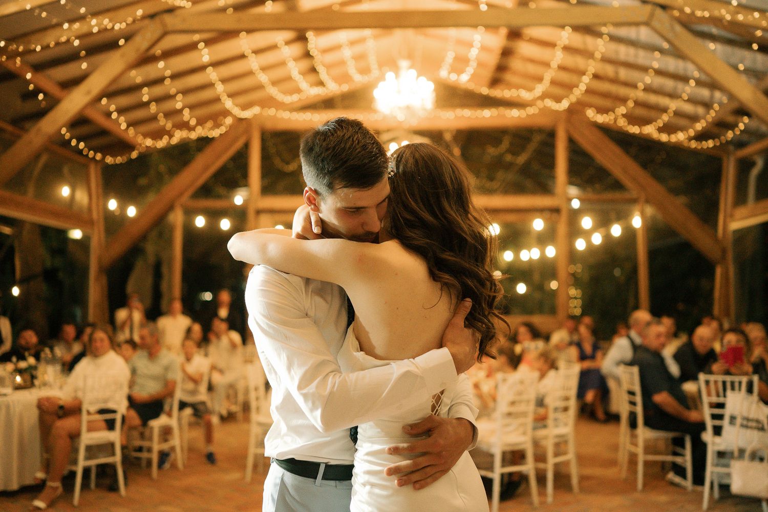A couple shares their first dance at a rustic barn wedding reception illuminated by romantic string lights.