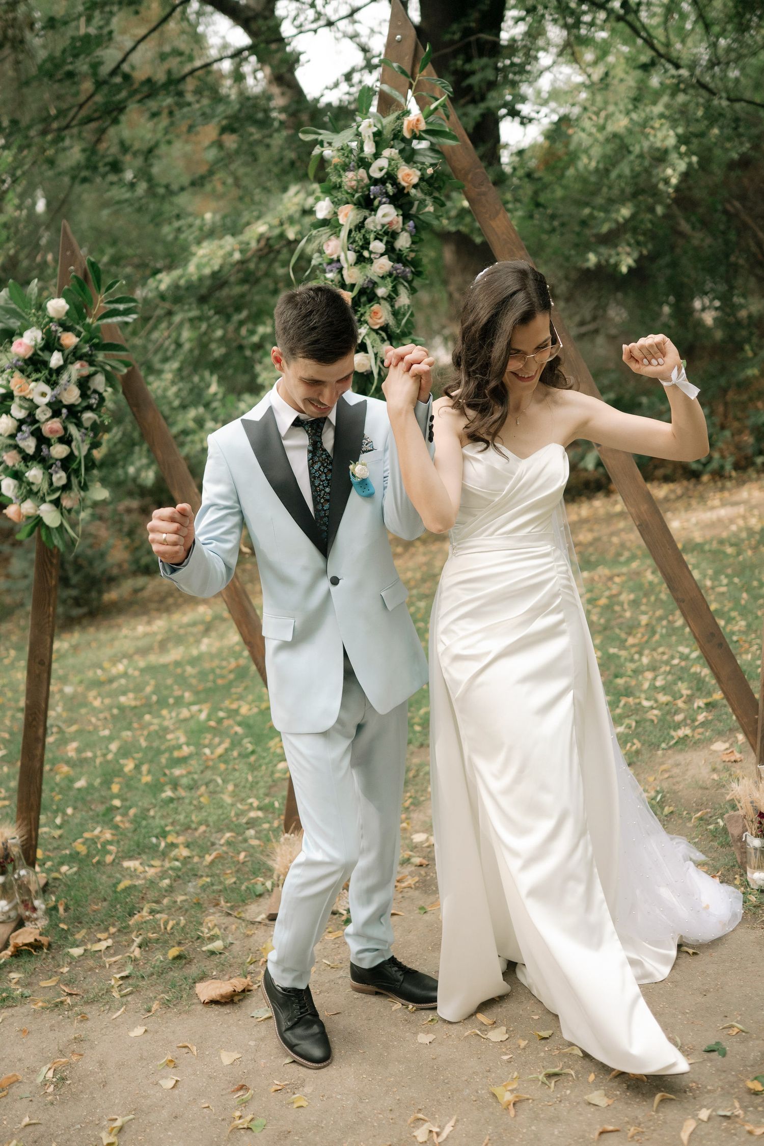 A couple dances playfully in a wooded outdoor setting with fallen autumn leaves.