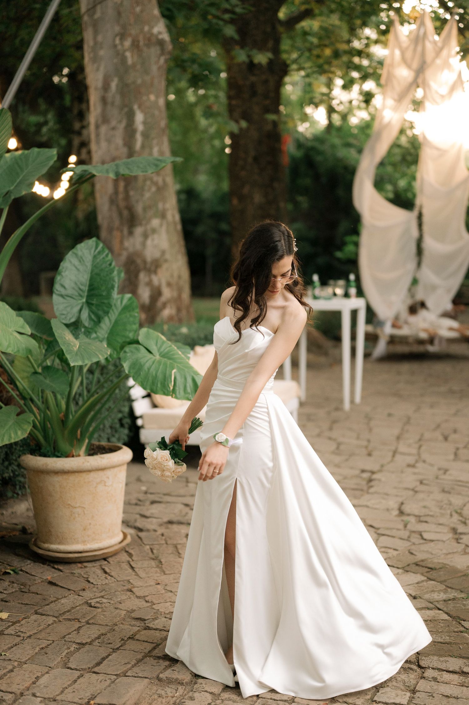 A bride in a white gown spins elegantly on a brick patio with lush greenery and string lights in the background.
