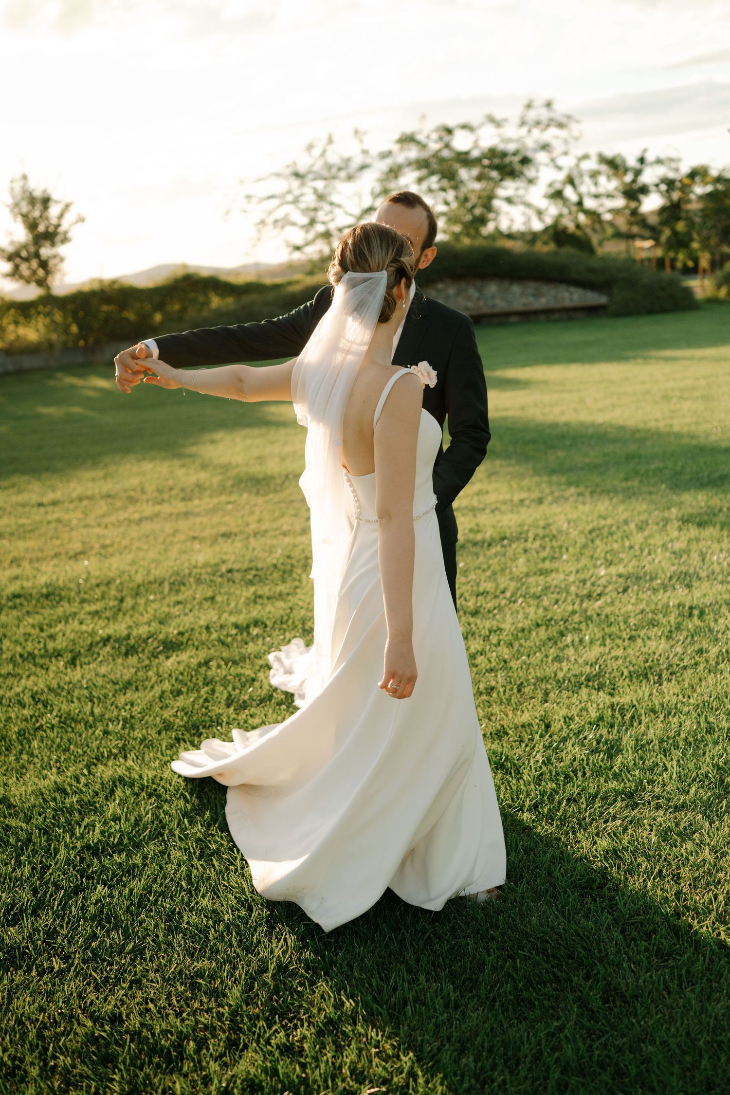 A couple in wedding attire dances together on a sunlit grassy hill during golden hour.