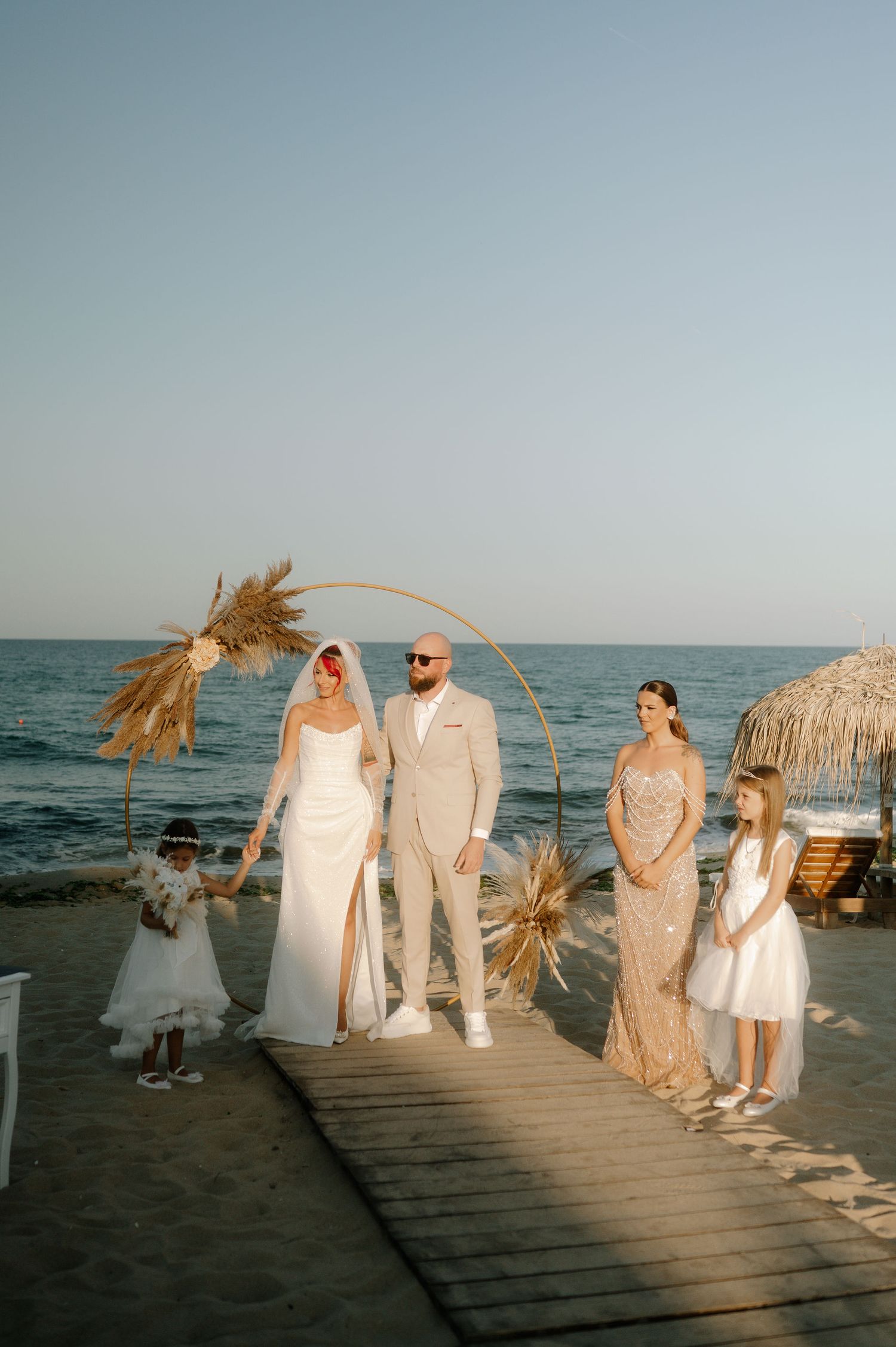 A beachside wedding ceremony on a wooden boardwalk with palm fronds and ocean views at sunset.