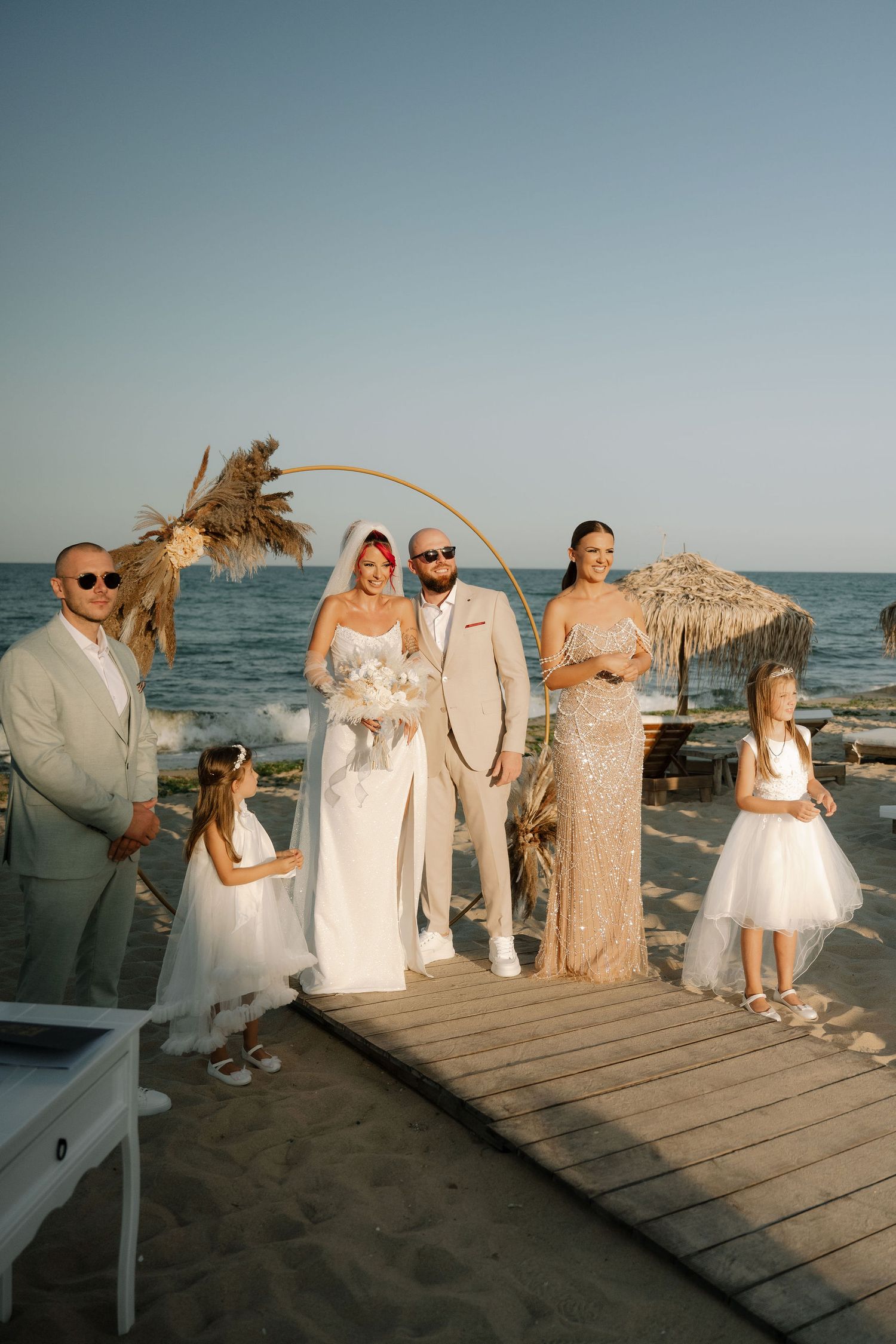 Wedding party poses on wooden boardwalk at beach during sunset with tropical palm trees and tiki huts in background.