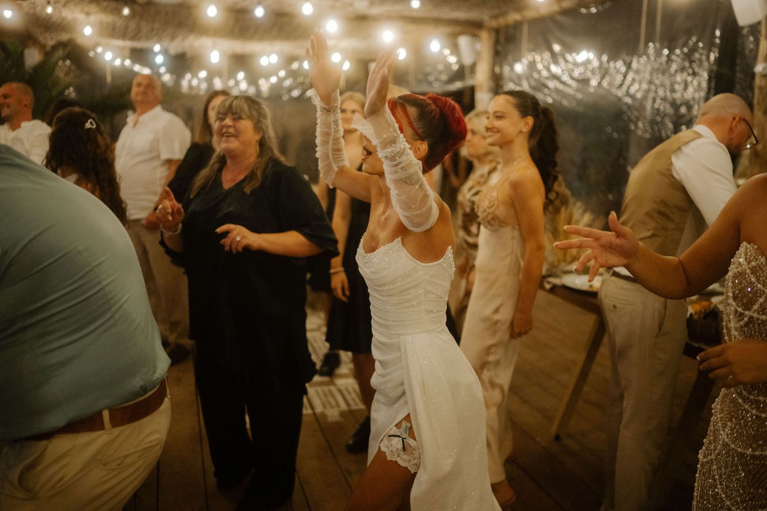 Wedding guests dance and celebrate together on a dimly lit dance floor decorated with string lights during a reception.