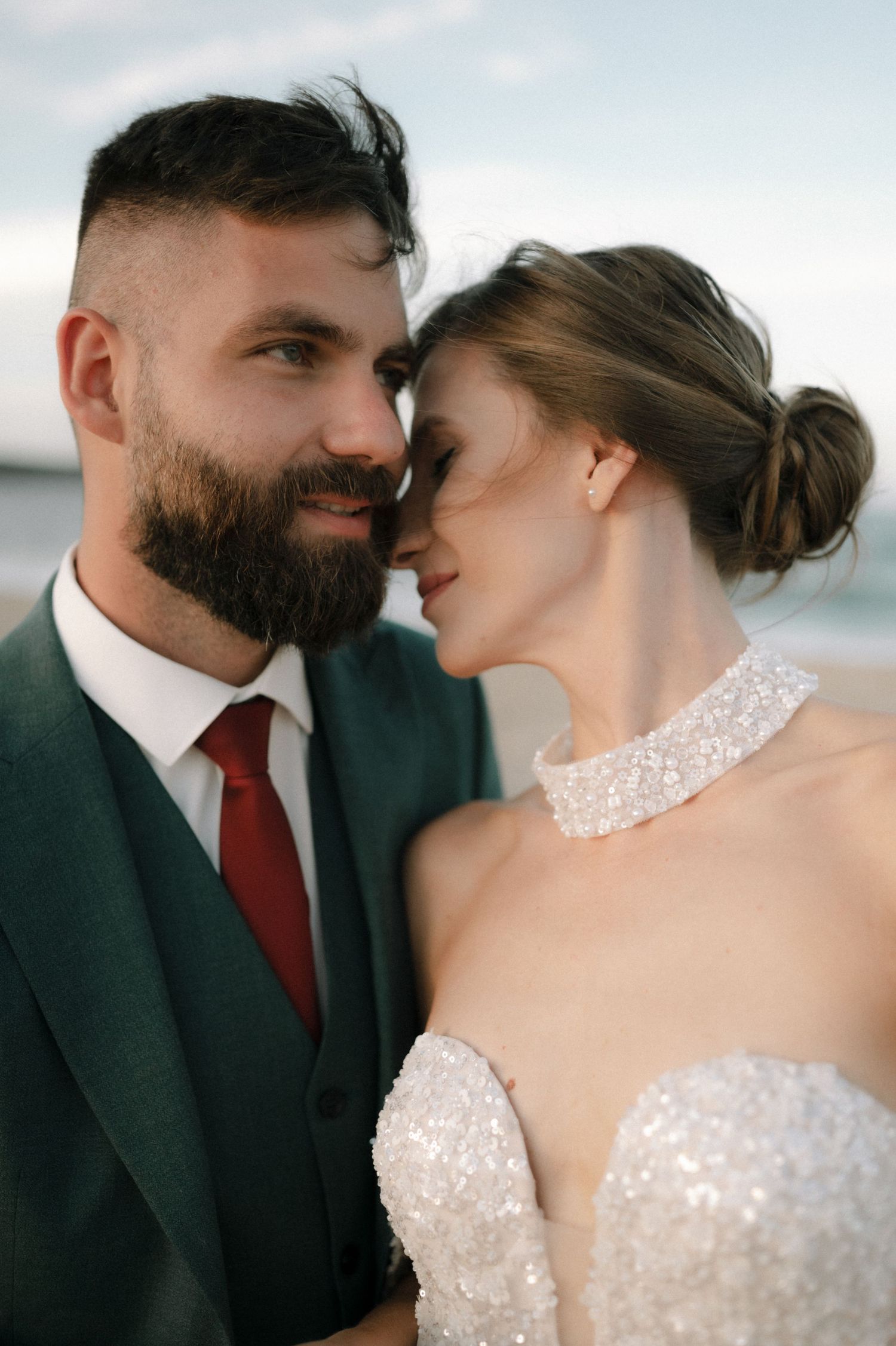 A bearded groom in a green suit with red tie stands close to his bride in a beaded gown on a beach.