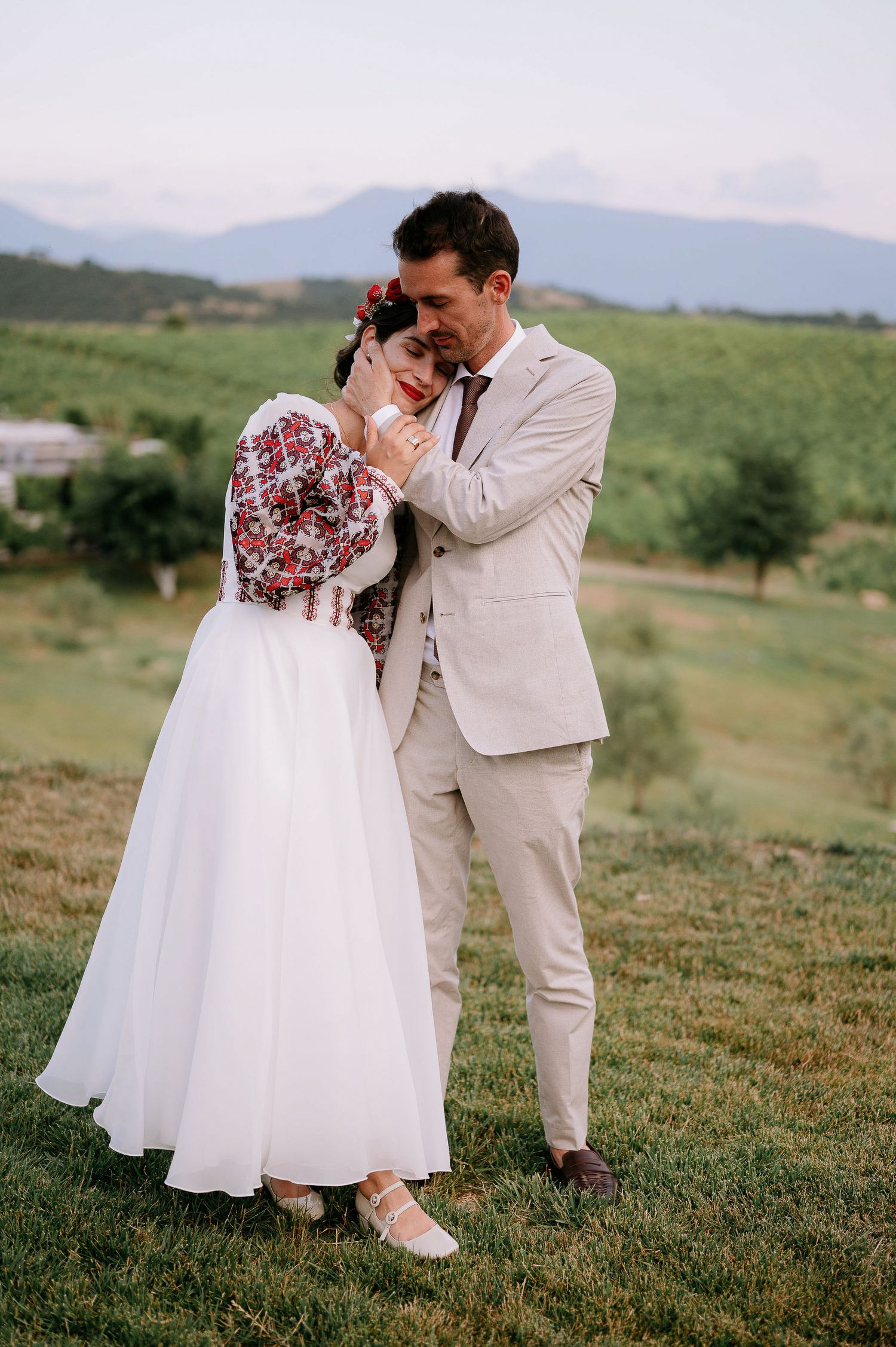 Tender moment between newlyweds on grassy hillside wearing traditional dress and suit.