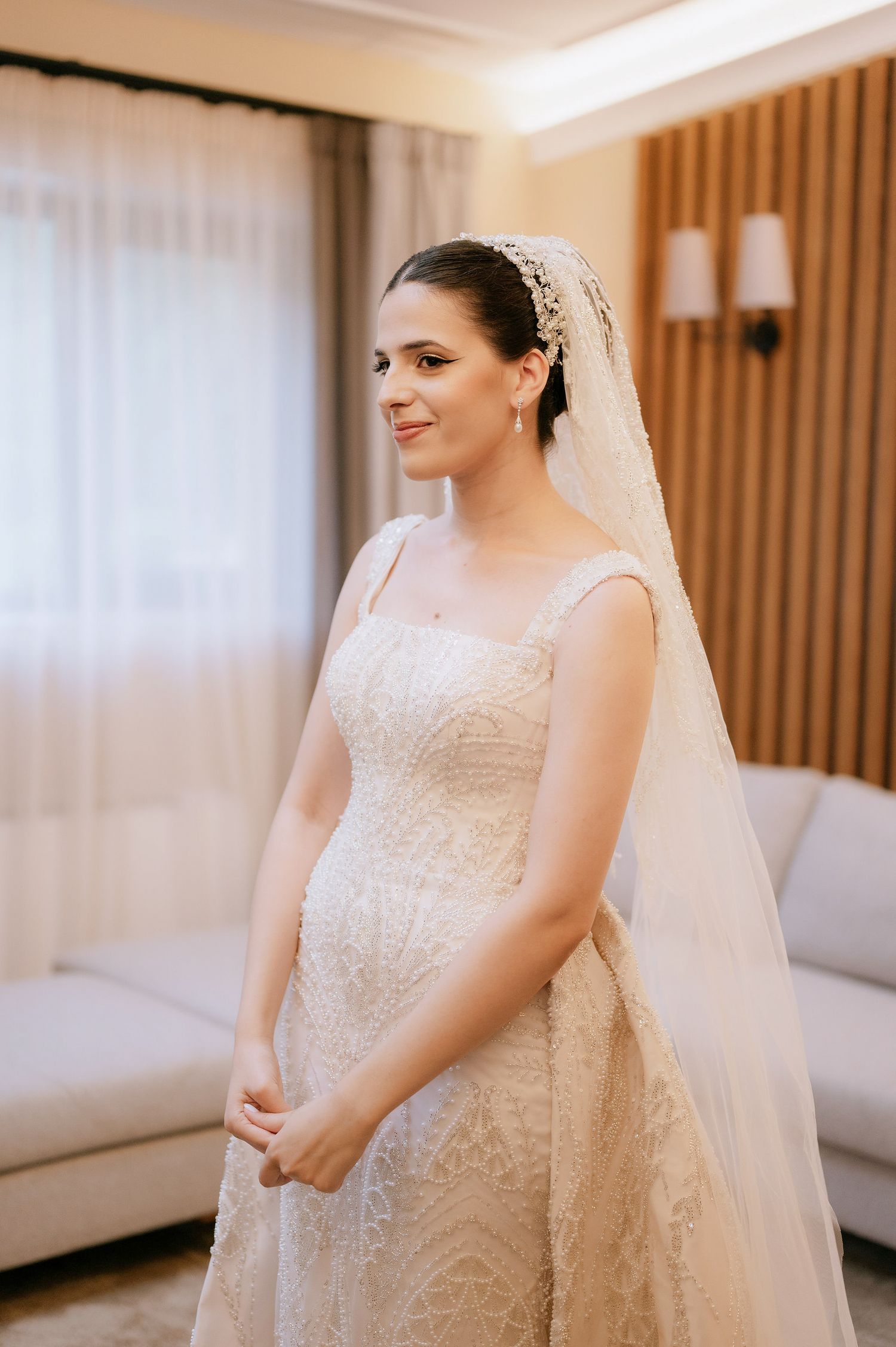 Elegant bride in a fitted lace wedding dress and long veil poses in front of wooden accent wall.