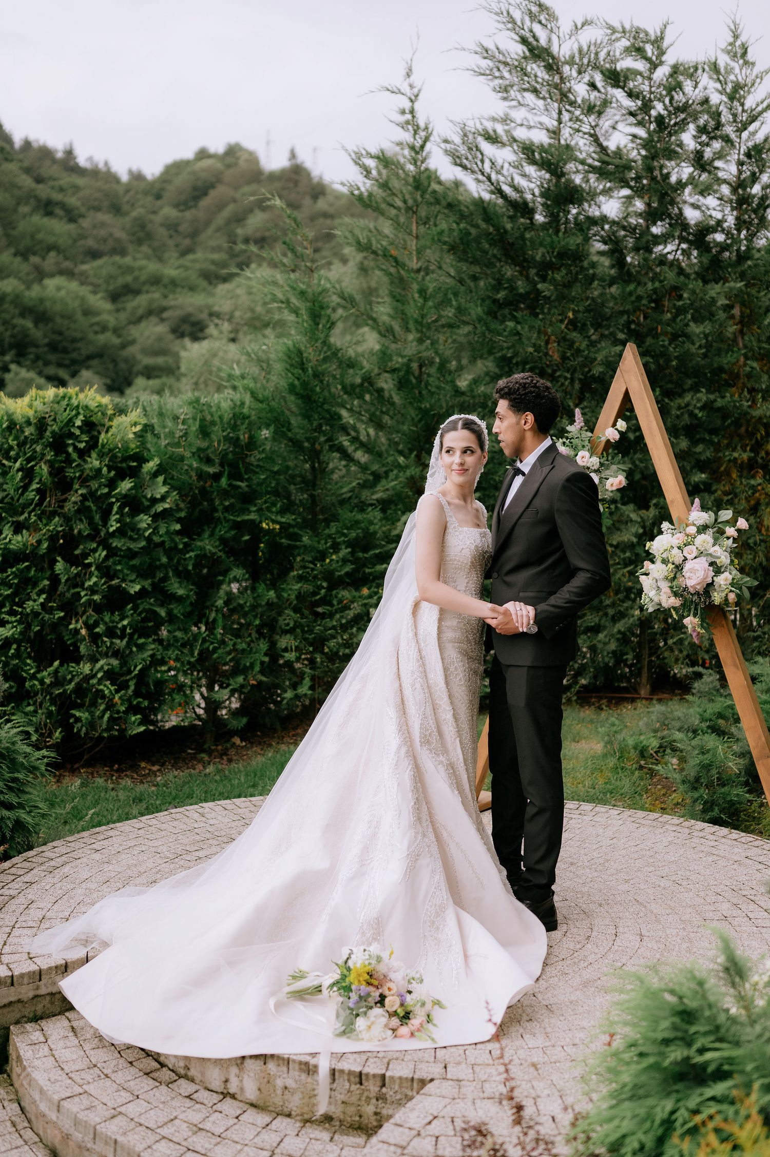 Elegant wedding portraits on a circular stone patio with a wooden triangle arch and evergreen backdrop.
