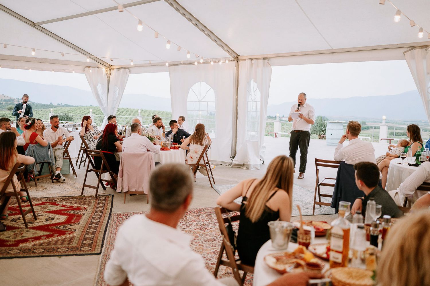 Wedding guests gather under an elegant white tent reception with mountain views and decorative rugs.