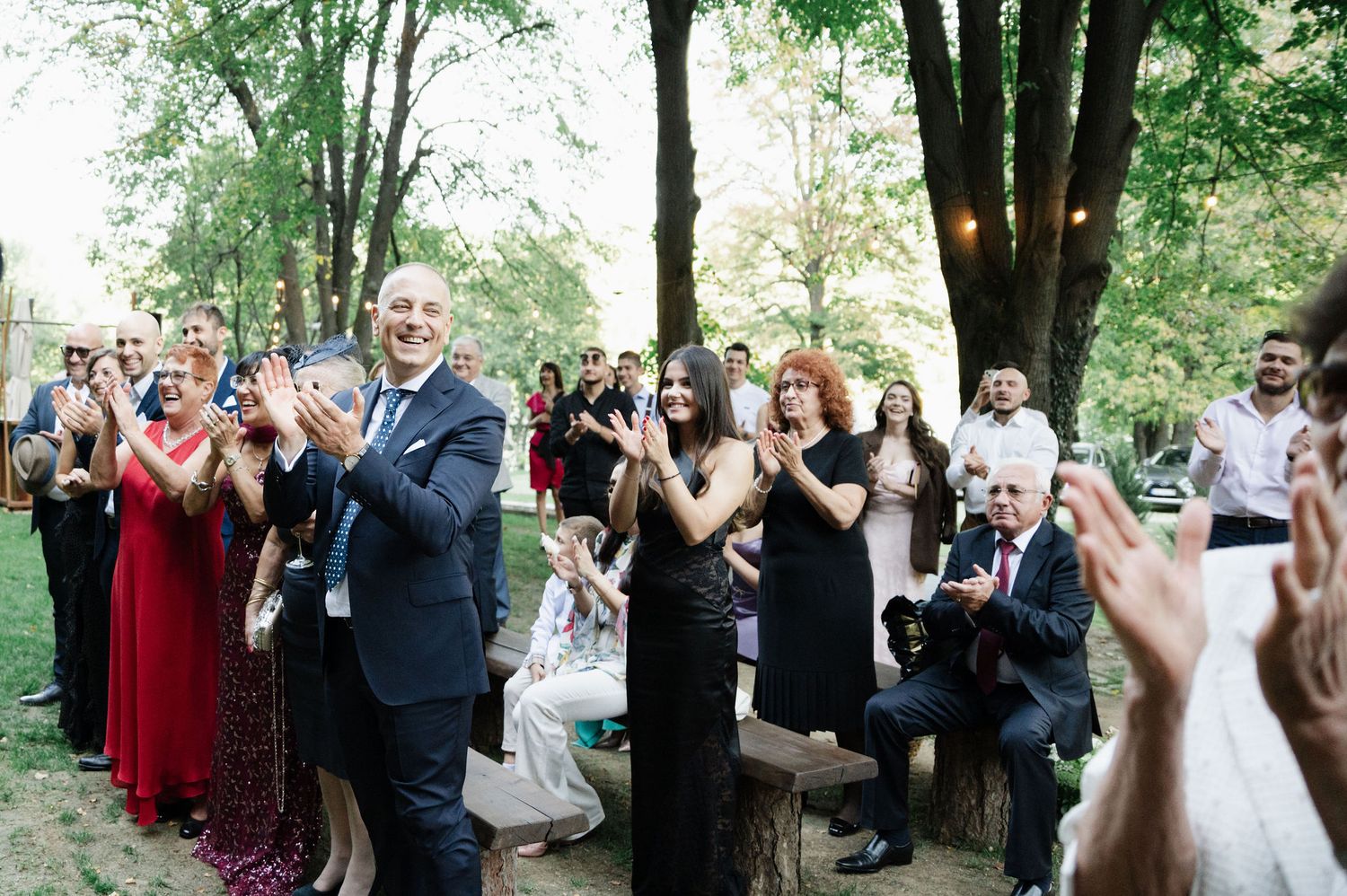 Wedding guests applaud during outdoor ceremony surrounded by trees.