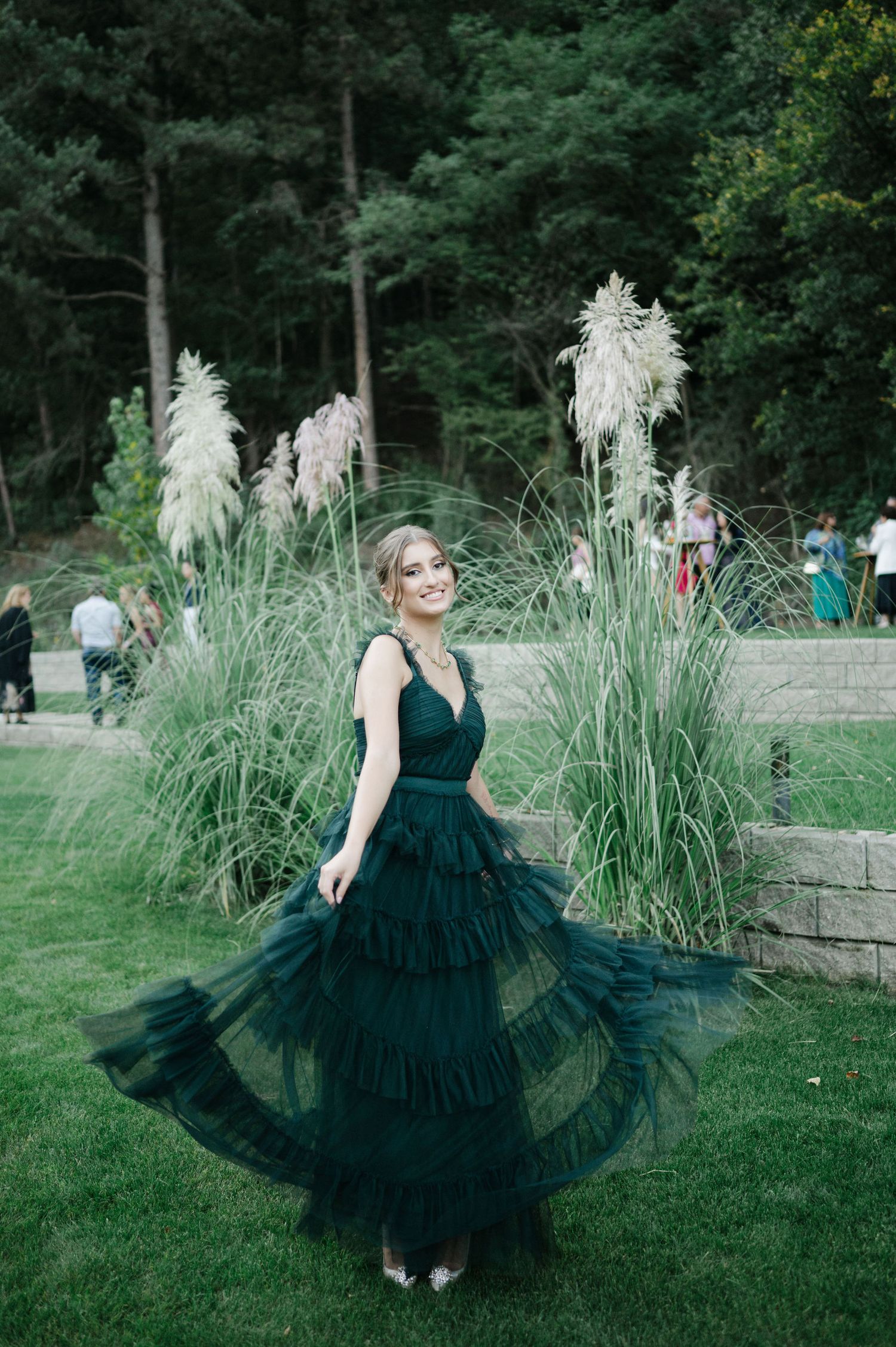 A person wearing a flowing dark green gown spins elegantly on a grassy lawn beside tall pampas grass.