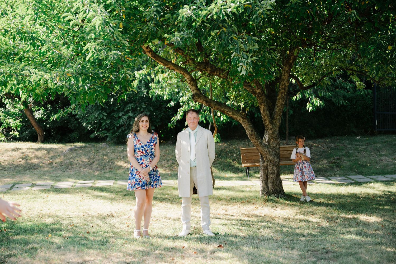 People gather under trees in a park-like setting on a sunny day.